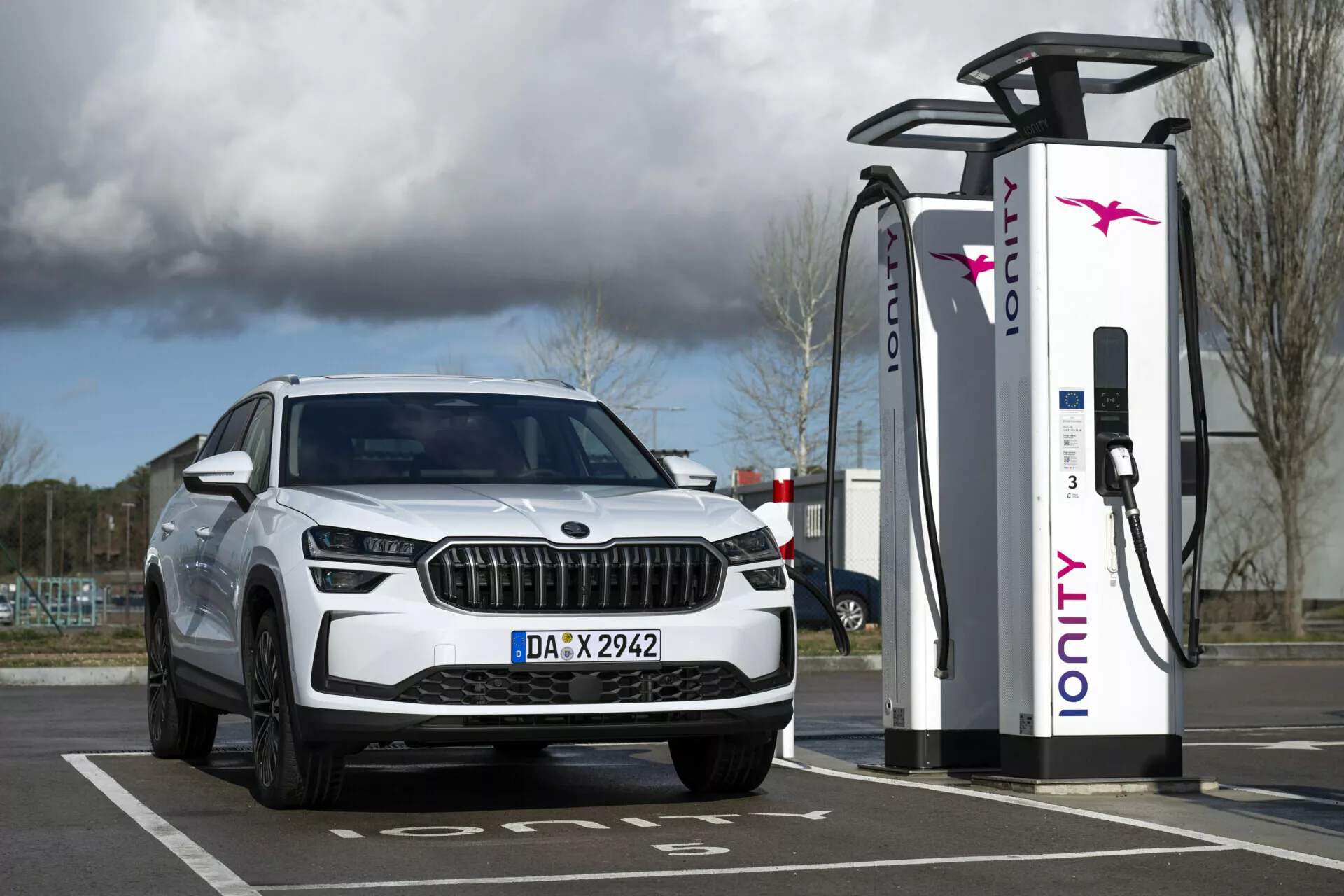 SUV bianco Skoda Kodiaq 2024 parcheggiato presso una stazione di ricarica elettrica, con vista del quarto anteriore dotato di fari LED e luci diurne, che sottolineano le sue capacità elettriche o ibride e il design lussuoso della griglia.