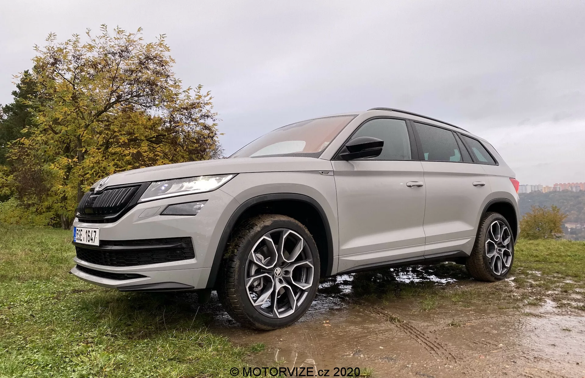 Vista frontale di tre quarti di un SUV Škoda Kodiaq 2017 presa da un'angolazione leggermente bassa, enfatizzandone l'altezza e la postura. Il veicolo è parcheggiato su una superficie irregolare di erba e fango, evidenziando le sue capacità fuoristrada. Lo Škoda Kodiaq presenta una griglia prominente e fari LED affilati. Il design meticoloso include grandi ruote in lega multirazza, un'elevata altezza da terra e pneumatici per tutte le stagioni. La carrozzeria è dipinta in un colore metallico neutro con vetri oscurati, e gli accenti cromati ne migliorano l'appeal premium. Le funzionalità moderne sono evidenti negli specchietti laterali in tinta carrozzeria con indicatori di direzione integrati e possibilmente sistemi di avviso dell'angolo cieco.
