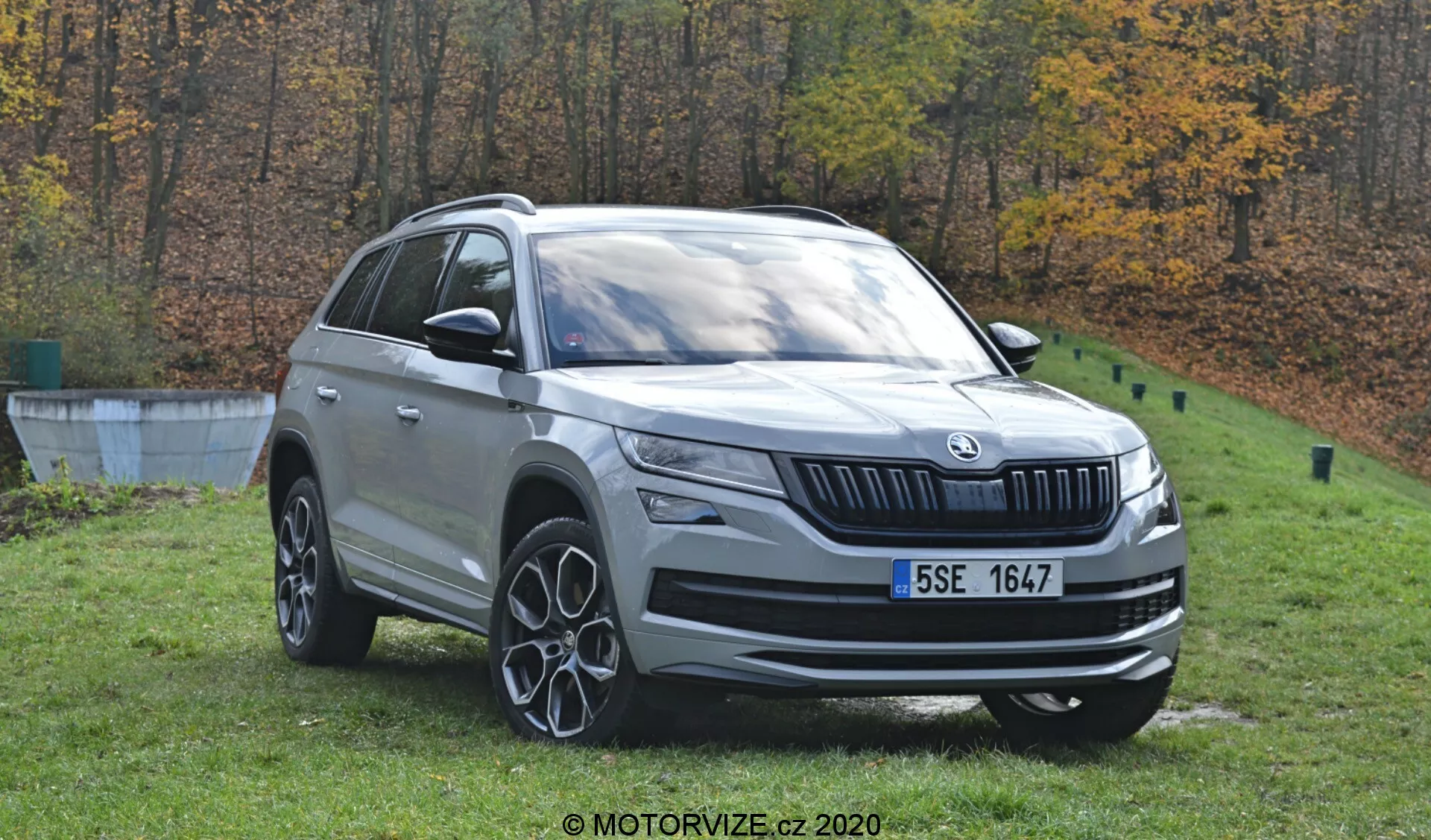 Una vista dettagliata della Škoda Kodiaq 2017, un SUV color argento, posizionato all'aperto su uno sfondo autunnale di alberi. L'immagine diurna cattura il veicolo da un angolo di tre quarti frontale, mostrando il profilo anteriore e laterale. Sono visibili elementi di design distintivi, tra cui l'emblema del marchio sulla griglia anteriore, fari a LED, fari fendinebbia, ruote in lega multispeziata, barre sul tetto e specchietti laterali con indicatori di direzione integrati. L'auto è posizionata sull'erba vicino a un bordo di cemento, suggerendo un ambiente simile a un parco.