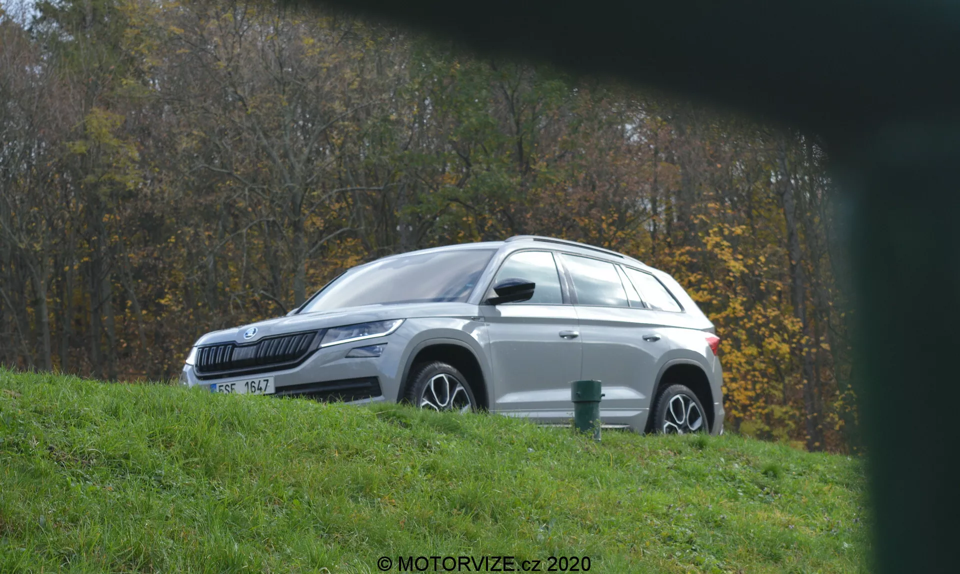 Vista laterale di un SUV Skoda Kodiaq bianco, probabilmente un modello moderno degli anni 2020, fotografato da una leggera angolazione elevata, parcheggiato su erba accanto a un sentiero asfaltato durante l'autunno, con cielo nuvoloso; che mostra porte chiuse, barre portatutto argento e fogliame autunnale sullo sfondo, e con una filigrana 'MOTORVIZE.cz 2020'.
