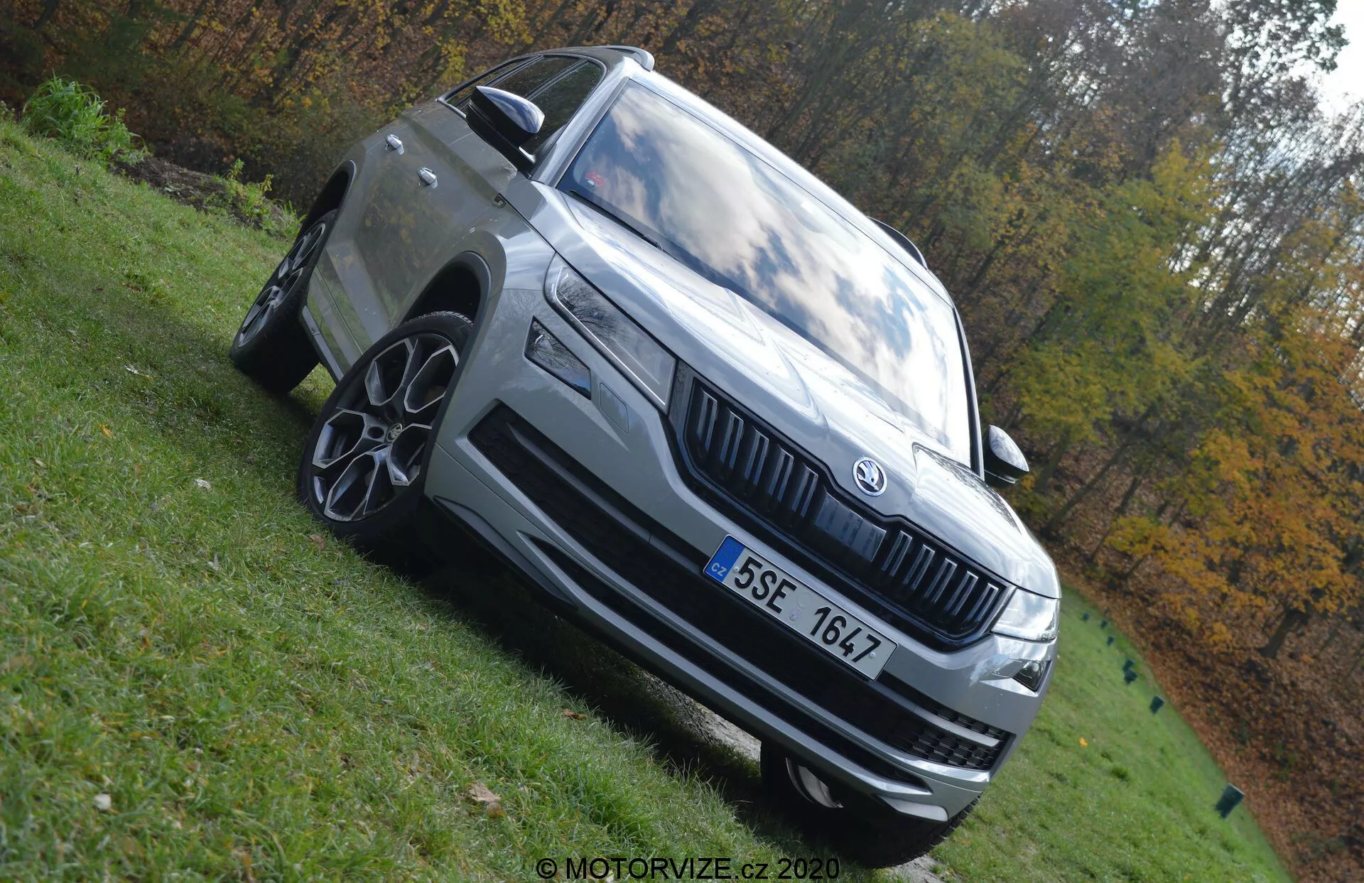 Fotografia a bassa angolazione di una Skoda Kodiaq 2017 che evidenzia la sua statura dominante. L'auto è rappresentata mostrando il quarto anteriore sinistro con caratteristiche notevoli come fari a LED, griglia prominente con emblema Skoda, cerchi in lega multisprort, paraurti anteriore con prese d'aria, specchietto laterale con indicatore integrato e linee di design esterne distintive. Il veicolo è di colore grigio chiaro o argento ed è parcheggiato su un'area erbosa sotto la luce naturale del giorno. Lo sfondo autunnale sfocato accentua la presenza del veicolo.