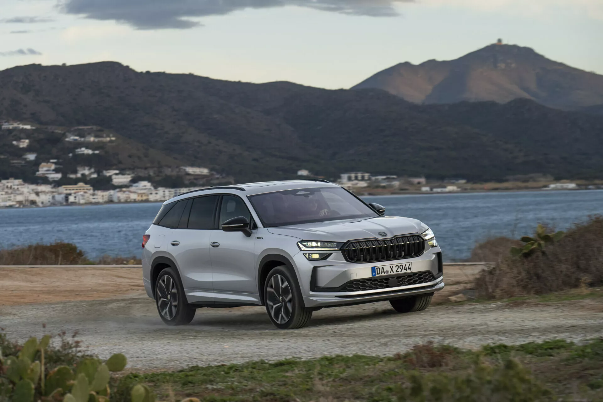 SUV Skoda Kodiaq 2024 parcheggiata su una superficie non asfaltata, con sfondo di colline sceniche e acqua, evidenziando i fari moderni a LED del veicolo, la griglia anteriore con possibile integrazione ADAS e il design contemporaneo delle ruote sotto una luce diurna morbida e nuvolosa.