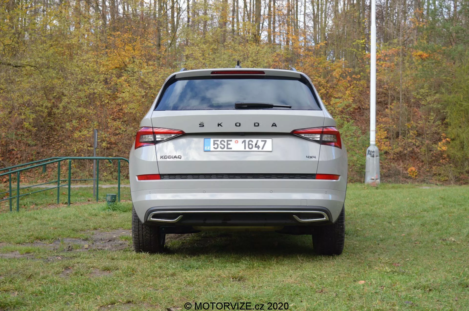 Immagine del retro di un veicolo Škoda Kodiaq 2017 in un contesto all'aperto. La scritta 'ŠKODA' e il nome del modello 'KODIAQ' sono ben visibili sul cofano del bagagliaio. Targa in stile europeo, luci posteriori moderne con luce antinebbia posteriore e luci di retromarcia integrate, paraurti posteriore visibile con sensori di parcheggio e parabrezza posteriore con tergicristallo sono chiaramente visibili. Gli elementi riflettenti nella parte inferiore del paraurti migliorano la visibilità. L'auto sembra parcheggiata su un'area erbosa e leggermente fangosa, suggerendo capacità off-road.