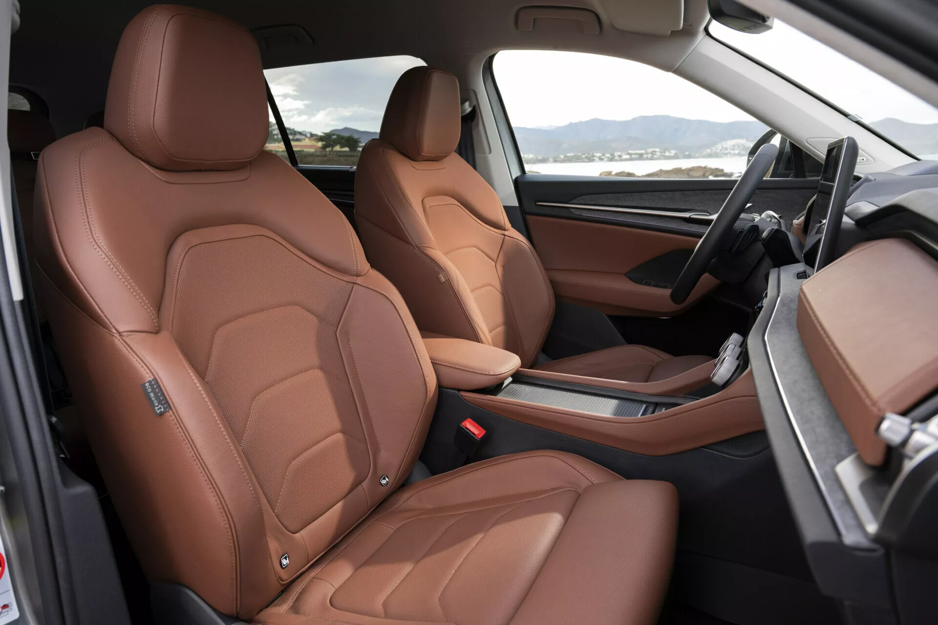 Vista interna della Škoda Kodiaq 2024 evidenziando sedili anteriori in lussuoso cuoio marrone con cuciture dettagliate, regolazione elettrica dei sedili e uno sguardo agli elementi di design di alta gamma come i rinforzi laterali ispirati allo sport, cruscotto bicolore nero e marrone, e console centrale minimalista con leva del cambio automatica, dall'apertura lato passeggero