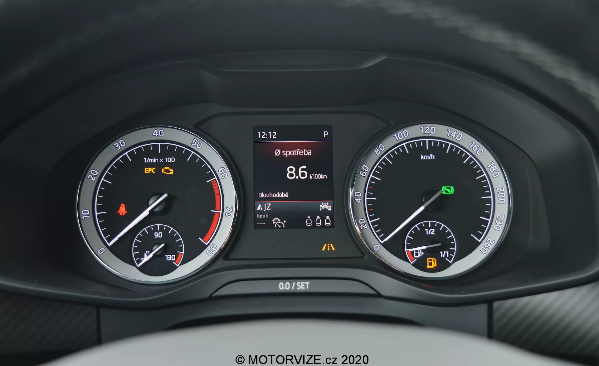 Vista dall'occhio del conducente del quadro strumenti della Škoda Kodiaq 2017, con un design moderno e pulito che combina display analogici e digitali. A sinistra: tachimetro analogico e indicatore del carburante, al centro: display multi-informazioni digitale che mostra l'ora (12:12), la marcia (P), la velocità (8.6 km/h), il contachilometri (15,666 km) e icone non determinate. A destra: tachimetro analogico con indicatore del livello di carica della batteria. Nel complesso, il cruscotto è prevalentemente scuro con illuminazione bianca.