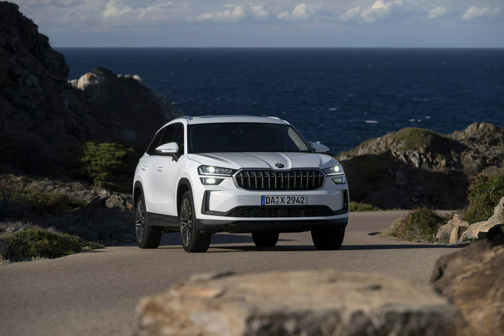 SUV bianco Skoda Kodiaq 2024 parcheggiato su una strada costiera con l'oceano sullo sfondo, caratterizzato da una prominente griglia frontale con listelli verticali cromati, luci diurne a LED, ruote in lega a più razze e una vista dettagliata dei tre quarti anteriori