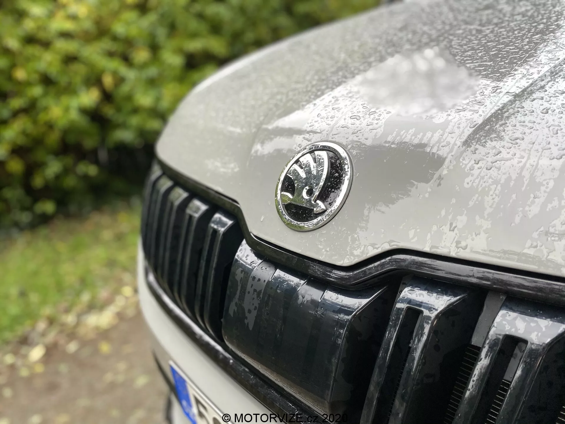Immagine ravvicinata della sezione anteriore di una Skoda Kodiaq 2017, con un focus particolare sulla griglia e sull'emblema del produttore. L'emblema circolare è posizionato al centro, mostrando dettagli delicati con gocce d'acqua che indicano una recente pioggia. L'angolo leggermente decentrato verso l'alto cattura il cofano e le lamelle verticali nere riflettenti della griglia in un'inquadratura nitida con una ridotta profondità di campo.