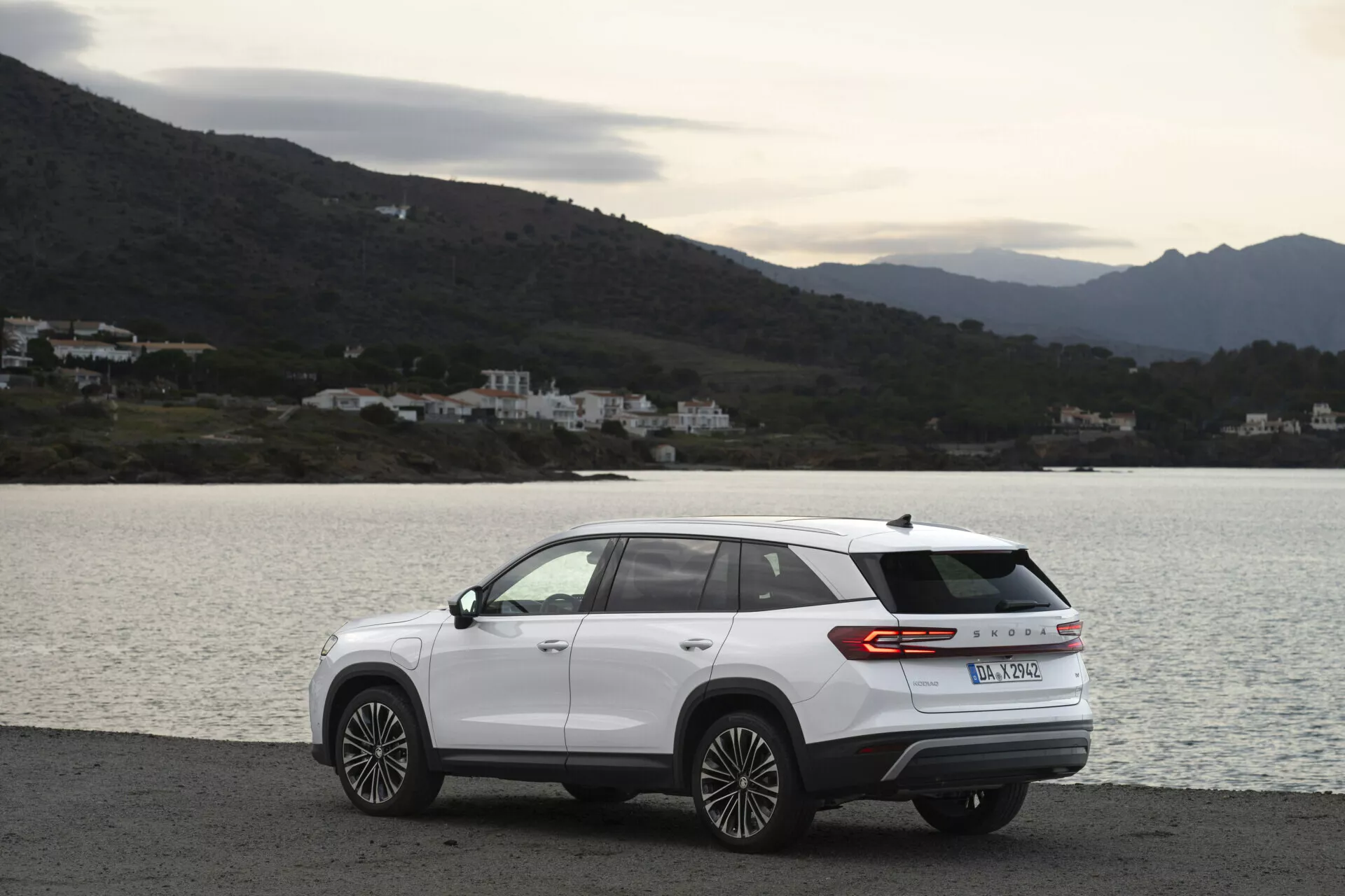 SUV moderno Skoda Kodiaq 2024 parcheggiato lateralmente su una strada costiera panoramica, con design aerodinamico, striscia di luce posteriore e ruote in lega a più razze, in una giornata nuvolosa con illuminazione naturale diffusa, con sfondo di acqua, colline e edifici