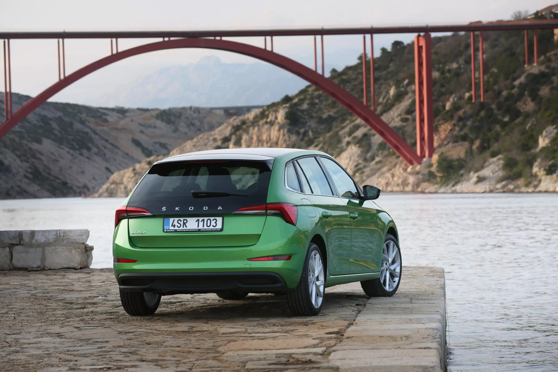 Skoda Scala verde del 2019 parcheggiata vicino all'acqua con vista del profilo posteriore e laterale, mostrando fanali posteriori, portellone, logo e designazione del modello su sfondo di un ponte rosso e montagne
