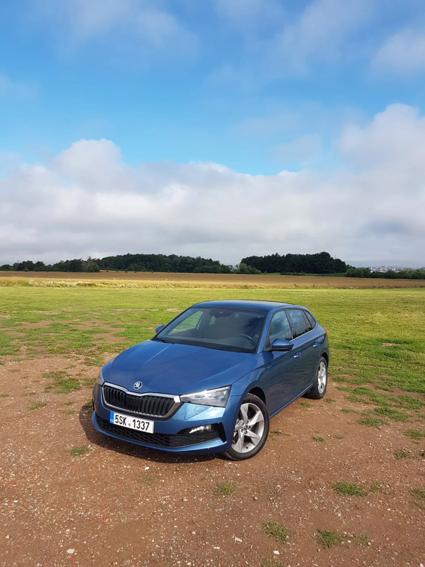 Berlina Skoda Scala 2019 in blu, parcheggiata sull'erba con cielo coperto, che mostra la griglia anteriore, i fari e il profilo del lato passeggero con sfondo di campagna