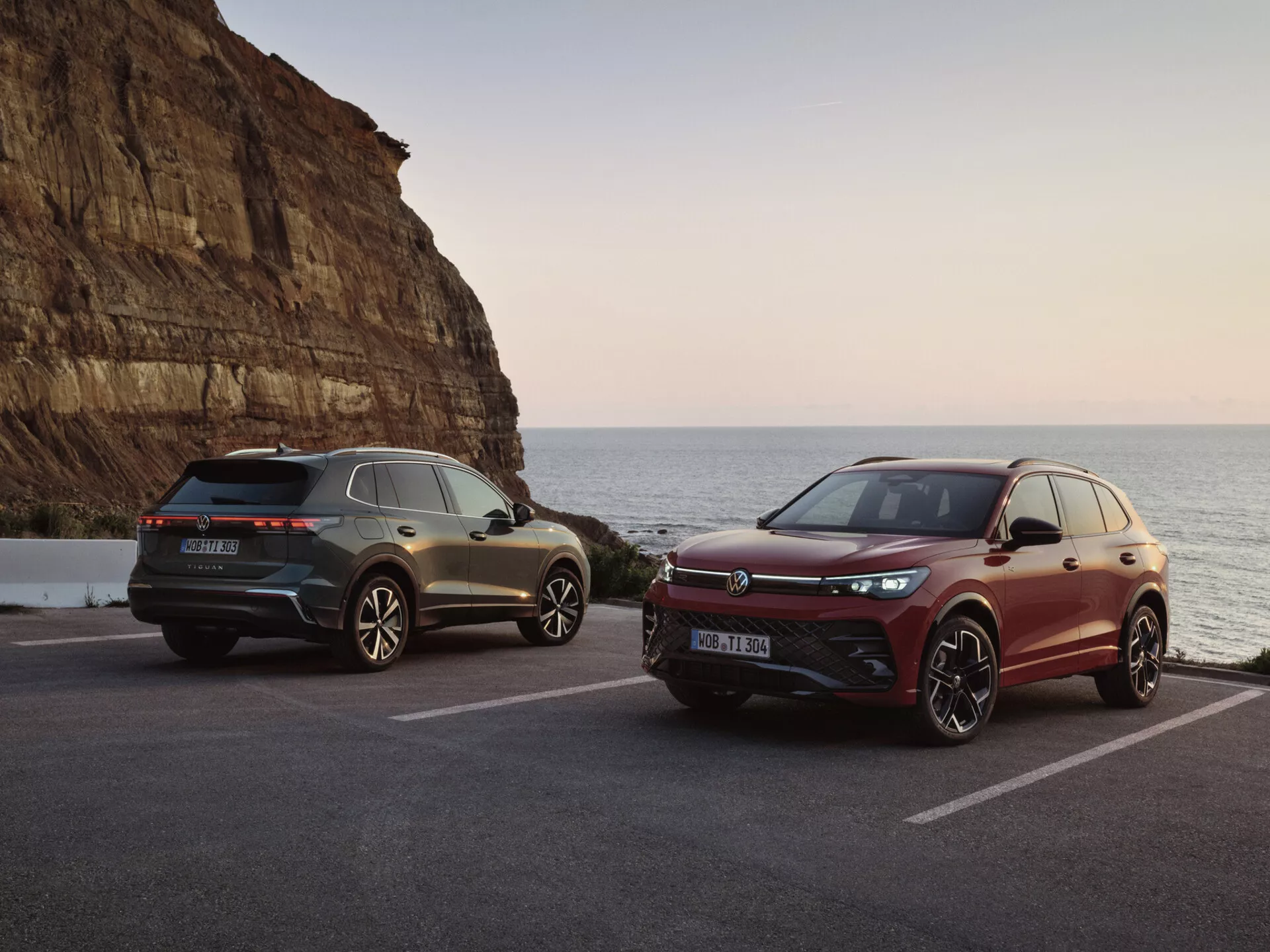 SUV di lusso in una scenografica ambientazione costiera al tramonto, caratterizzate da un modello grigio con fanali posteriori LED e elementi di design sportivo e un modello bordeaux con griglia frontale orientata alle prestazioni e sistemi avanzati di assistenza alla guida
