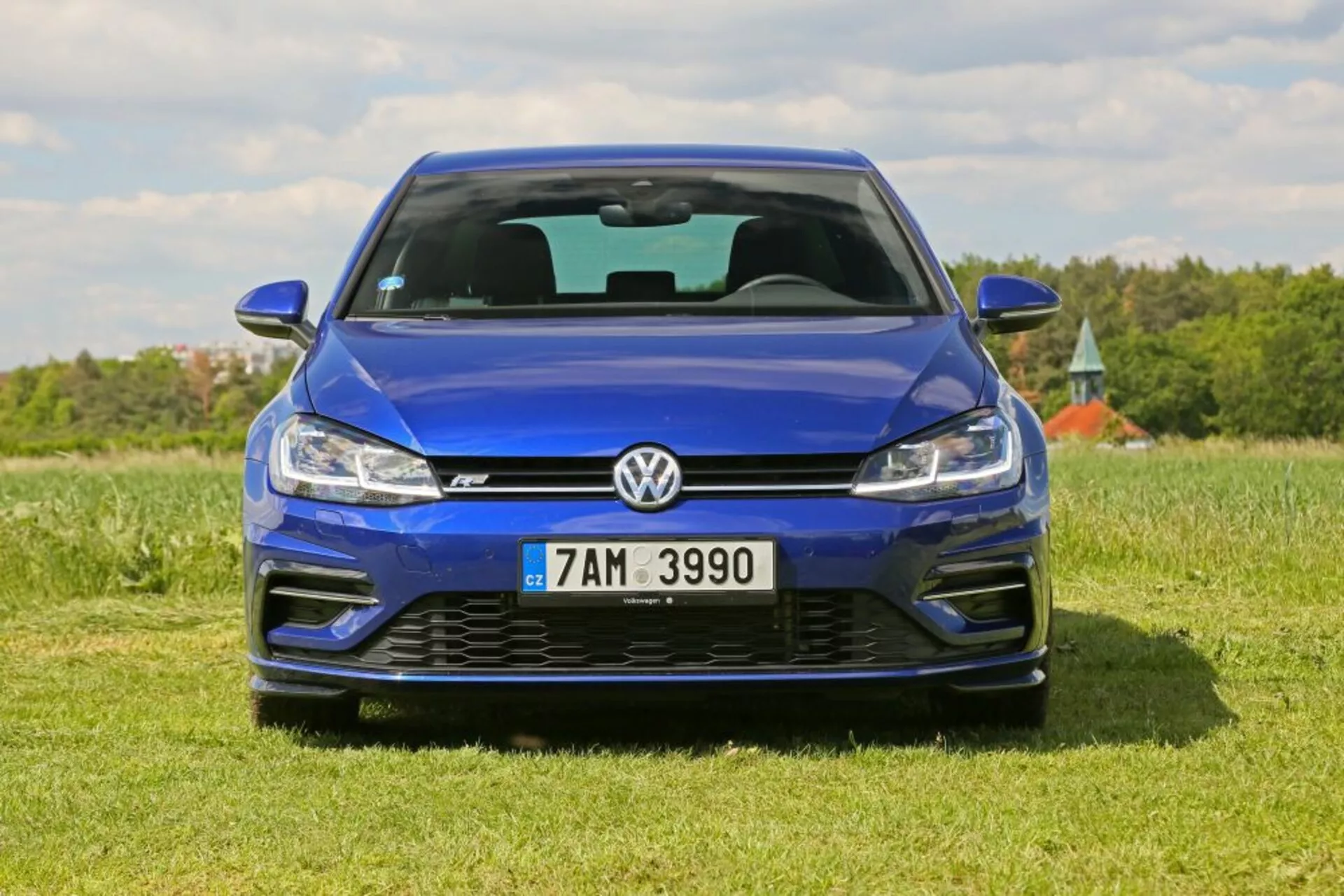 Veduta frontale di una Volkswagen Golf blu parcheggiata su un campo erboso sotto un cielo chiaro, mettendo in evidenza la sua finitura metallica, il design della griglia, la tecnologia dei fari LED e il paraurti anteriore sportivo inclusi gli aspiratori d'aria e l'emblema.