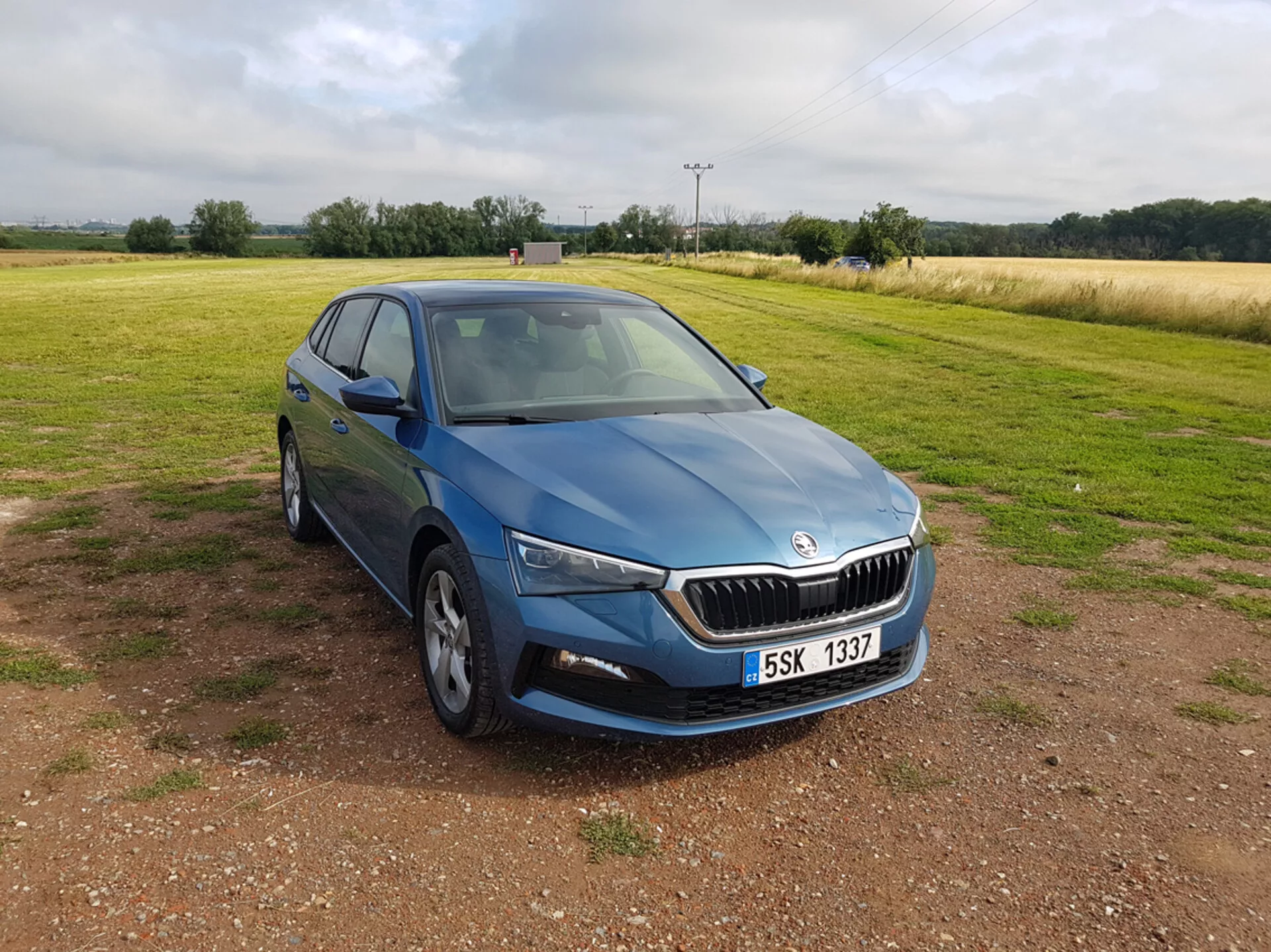 Skoda Scala blu 2019 vista da un angolo anteriore di tre quarti mettendo in evidenza design, fari LED, griglia cromata, ruote in lega a più razze, indicatori di direzione integrati negli specchietti laterali e finestre oscurate, posizionata su ghiaia con un campo sullo sfondo sotto cieli nuvolosi.