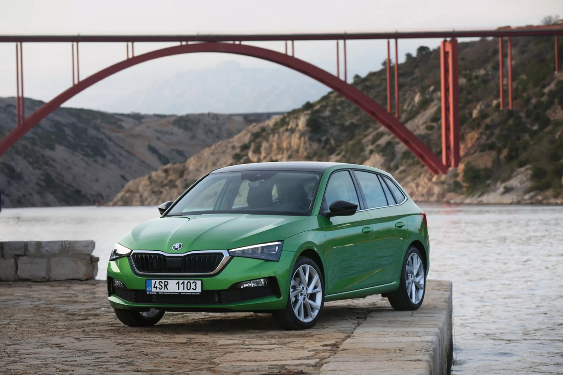 Hatchback verde a quattro porte Skoda Scala 2019 su molo in cemento vicino all'acqua con griglia visibile, fari LED, paraurti anteriore e ruote in lega, su sfondo di ponte scenico