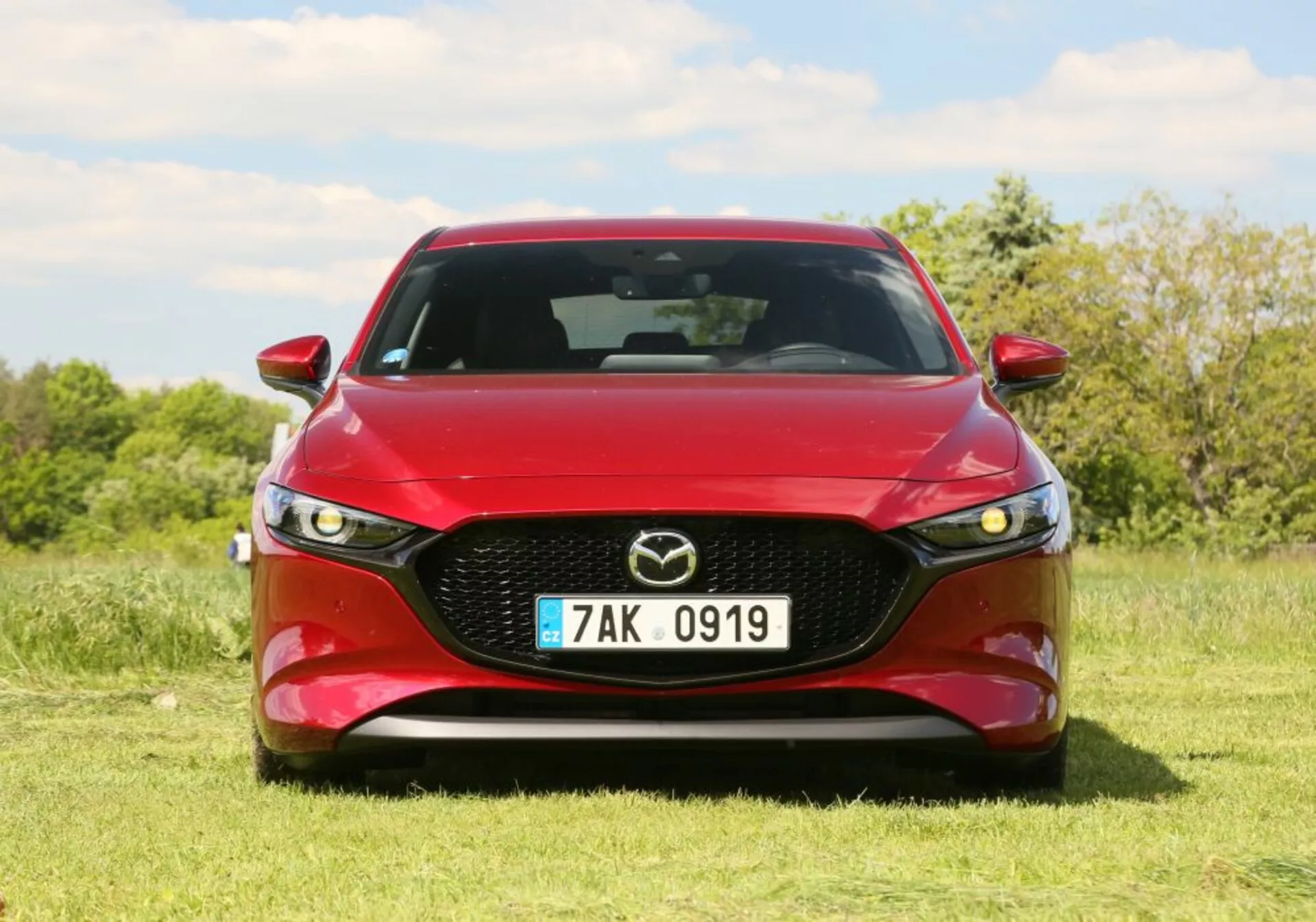 Veicolo Mazda rosso con fari LED e griglia cromata parcheggiato sull'erba sotto un cielo limpido, che mostra il design sportivo frontale ed estetica del veicolo.
