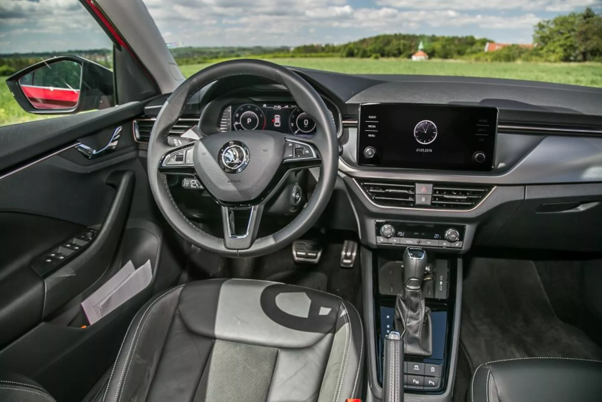Vista interna Skoda Scala 2019 dal lato passeggero anteriore con volante a tre razze con comandi integrati e quadro strumenti digitale, sistema di infotainment touch nella parte superiore della consolle centrale con controlli manuali del clima sottostanti, leva del cambio automatica, sedili anteriori profilati con rivestimento simil-pelle, pannello porta lato guidatore con regolazioni per finestrini e specchietti elettrici, illuminazione ambiente interna, specchietto retrovisore auto-oscurante e visibilità del paesaggio attraverso il parabrezza e la finestra lato guidatore, evidenziando il design ergonomico e la tecnologia integrata.
