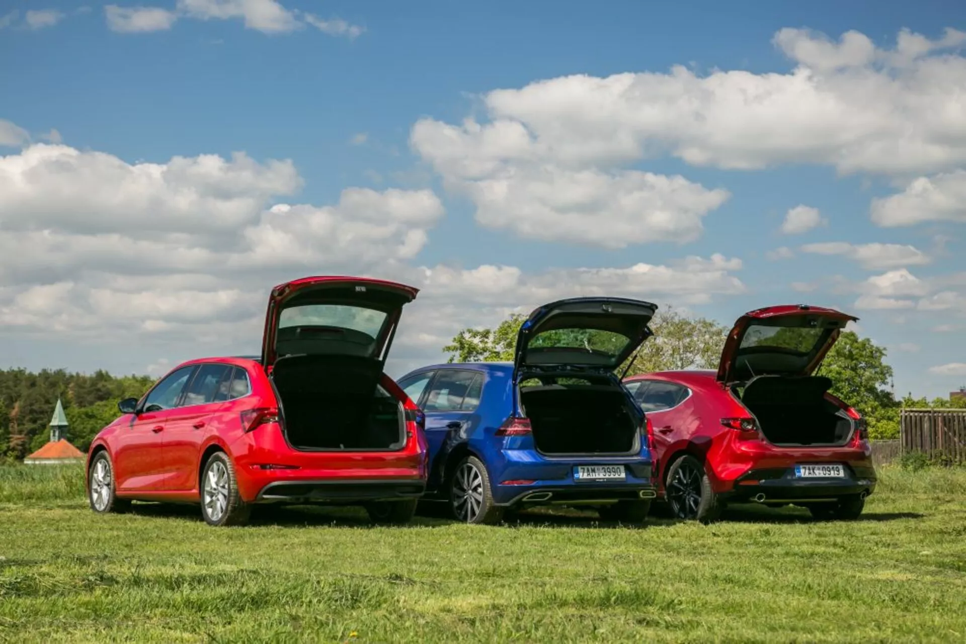 Modelli Skoda Scala 2019 in rosso e blu parcheggiati in un campo erboso, mostrano profili posteriori, bagagliai aperti per la funzionalità dello spazio di carico e dettagli di design sotto un cielo chiaro.