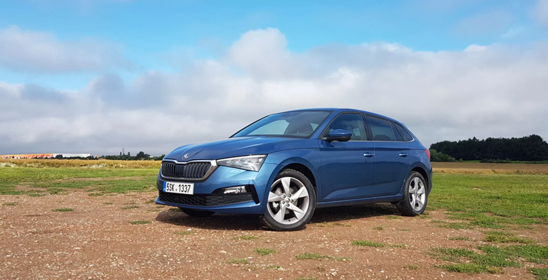 Skoda Scala hatchback blu 2019 su superficie non asfaltata dotata di una griglia frontale distintiva, fari moderni a LED, cerchi in lega e un elegante profilo laterale sinistro su sfondo di cielo parzialmente nuvoloso