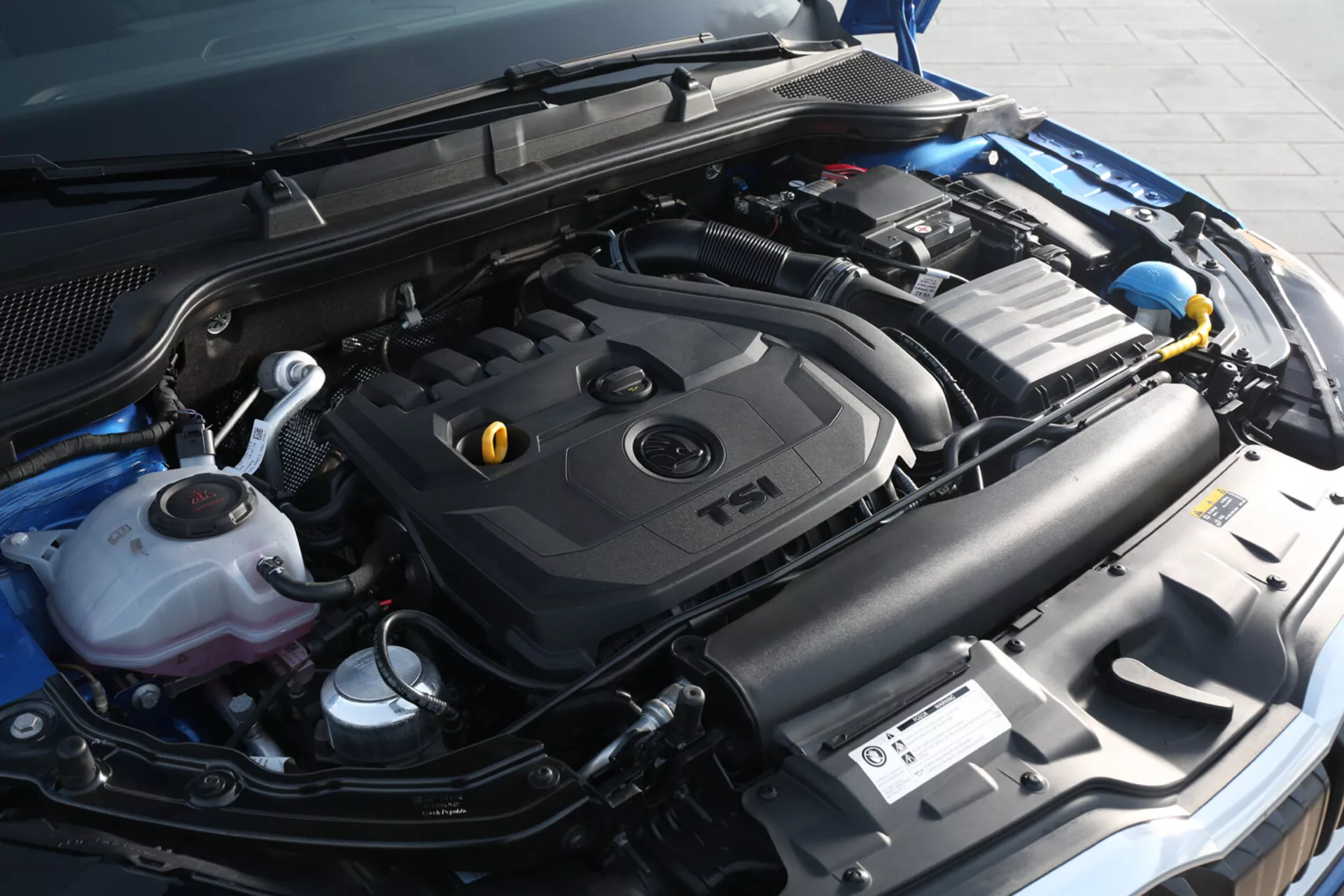Vista ravvicinata del vano motore di una Skoda Scala 2019, evidenziando il coperchio del motore TSI turbo e componenti inclusi il serbatoio del liquido di raffreddamento, contenitore del liquido lavavetri, batteria dell'auto e sistema di aspirazione da un angolo frontale leggermente a destra