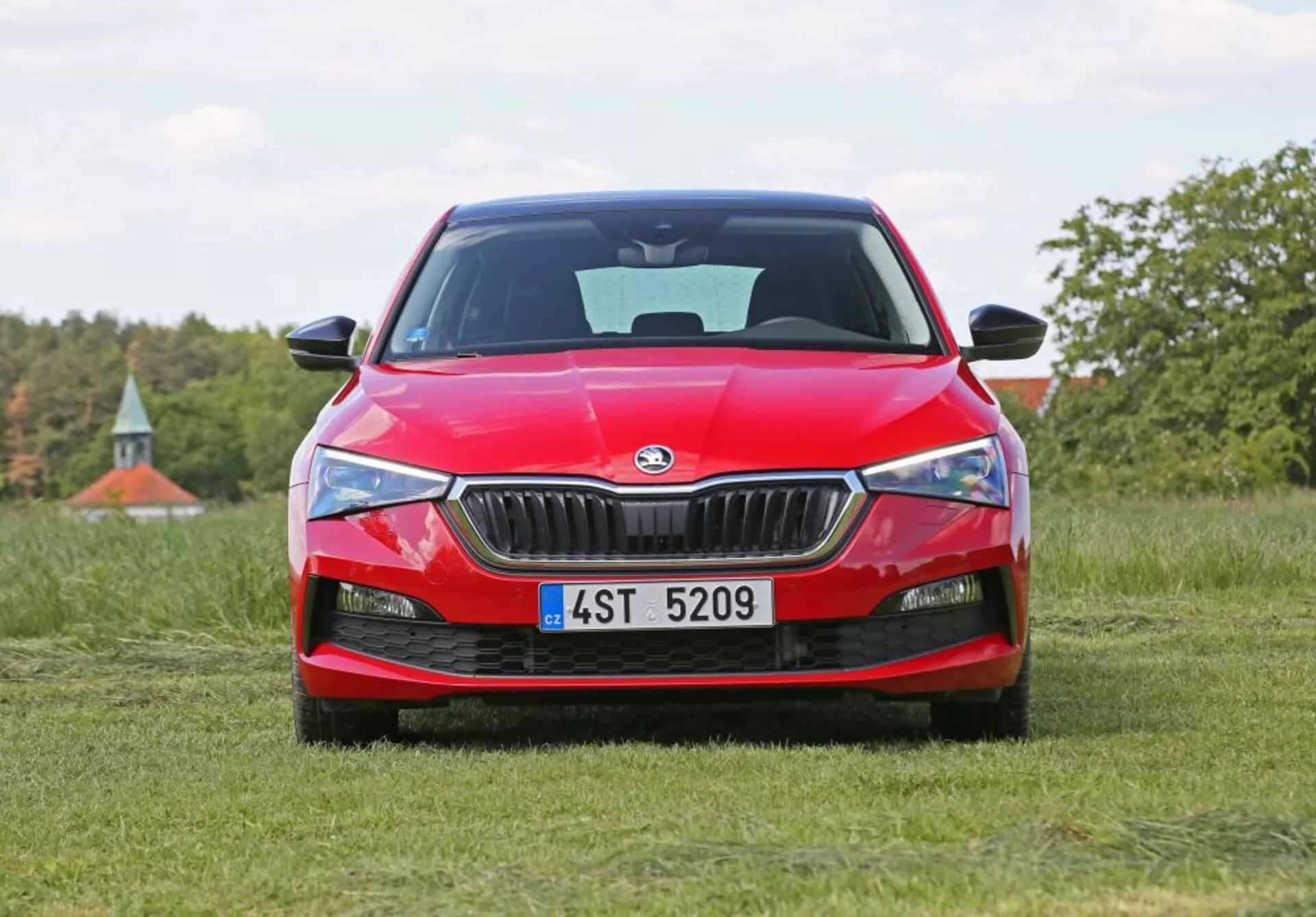 Skoda Scala 2019 di colore rosso posizionata su un campo erboso con vista frontale, mettendo in mostra la griglia frontale dell'auto, fari affilati e logo Skoda, su uno sfondo naturale con alberi e un accenno di cielo blu.