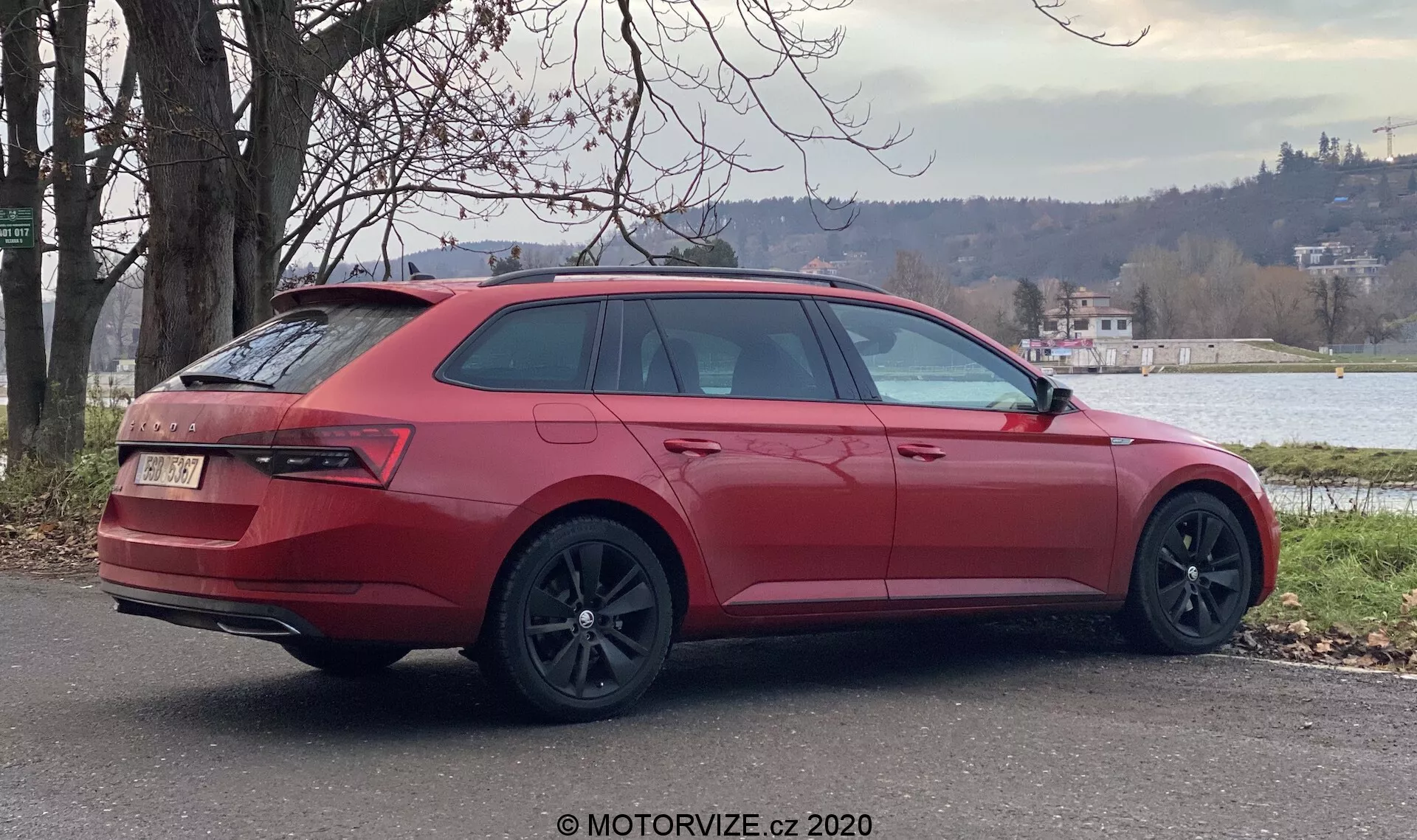 Una station wagon Skoda Superb 2019 rossa parcheggiata all'aperto, vista da un'angolazione posteriore di tre quarti che mostra il lato destro dell'auto. Gli elementi chiave del design includono grandi cerchi in lega scuri, vetri posteriori e laterali inclinati, fanali posteriori avvolgenti, portellone posteriore chiuso con stemma, paraurti posteriore con diffusore e doppi scarichi, barre sul tetto e specchietto laterale esterno abbinato al colore. L'ambiente presenta vegetazione e uno specchio d'acqua sullo sfondo, suggerendo un ambiente suburbano o semirurale tranquillo.