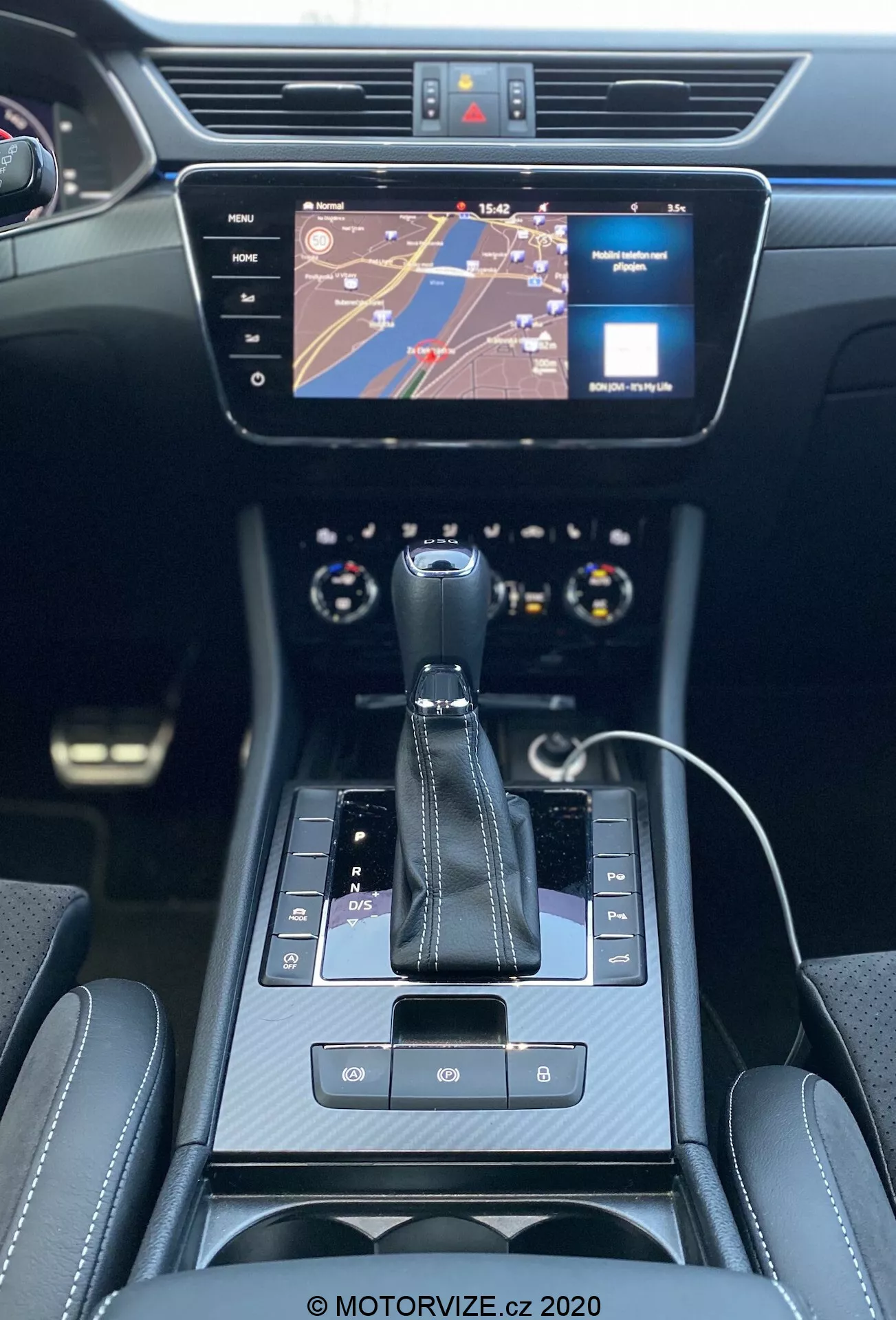 Vista interna della Skoda Superb Limousine 2019, incentrata sulla console centrale e sul cruscotto dalla prospettiva del sedile posteriore. Presenta una leva del cambio automatico con 'P' per il parcheggio visibile, pulsante del freno di stazionamento elettronico, pulsanti tattili e interfaccia touchpad. Un touchscreen di infotainment mostra una mappa e un indicatore multimediale. Controlli del clima a doppia zona sotto lo schermo, fiancheggiati da bocchette dell'aria. Interno moderno ed elegante in nero con finiture cromate e sedili lussuosi con cuciture visibili. La parte superiore del volante multifunzionale e parte del quadro strumenti con quadranti analogici sono visibili. Cavo di ricarica per smartphone che suggerisce funzionalità di connettività.