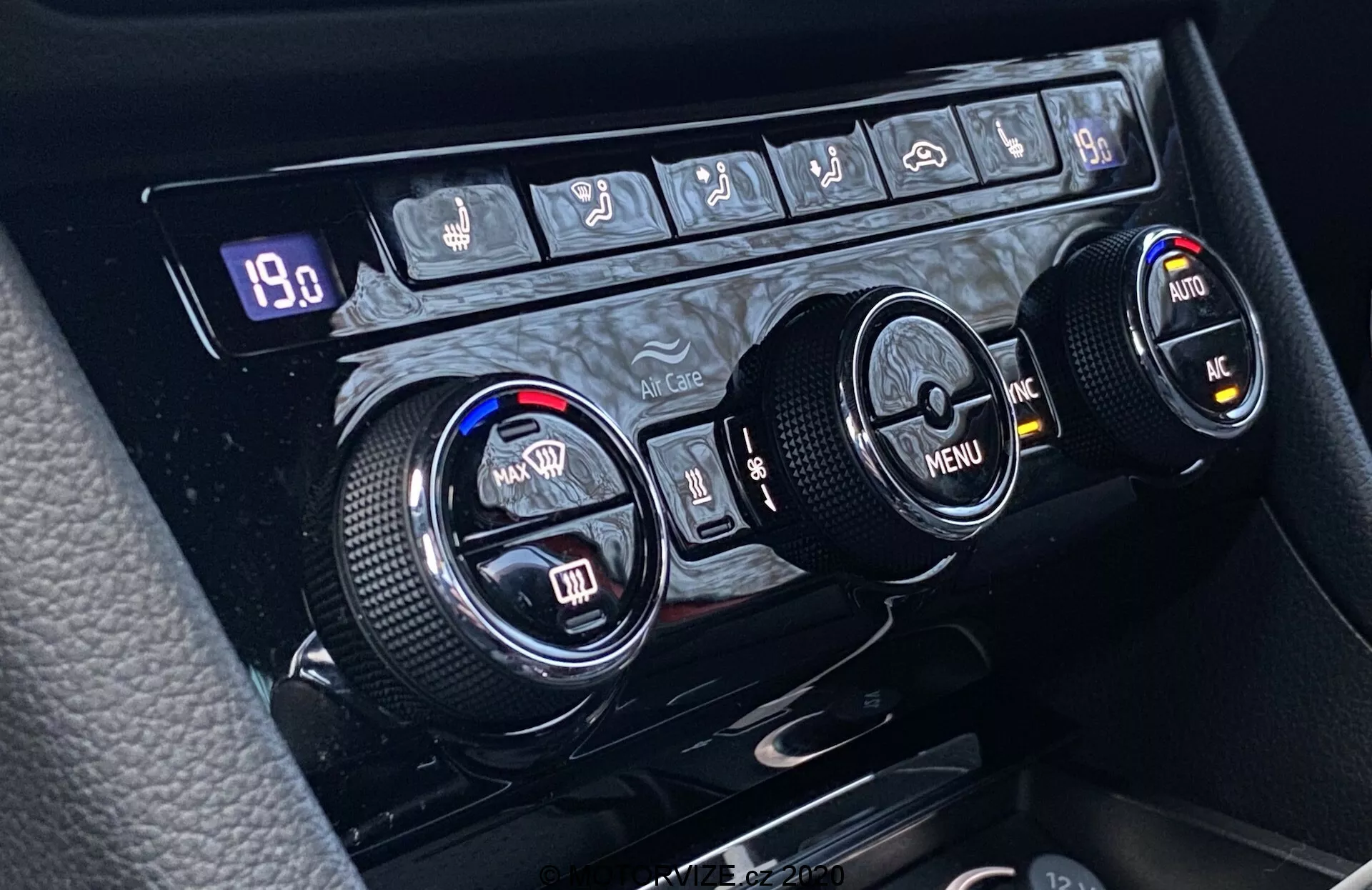 Primo piano del sistema di controllo del clima della Škoda Superb 2019 posizionato sulla console centrale, con tre manopole circolari per la regolazione della temperatura, la navigazione nel menu e funzioni aggiuntive. La manopola sinistra regola la temperatura per il conducente e quella destra per il passeggero, mentre la manopola centrale offre navigazione nel menu con frecce direzionali. I bordi testurizzati garantiscono un uso ergonomico. Sopra le manopole, una fila di pulsanti include controlli per il disappannamento del lunotto posteriore, il disappannamento del parabrezza anteriore, la recircolazione dell'aria, il controllo climatico automatico, l'aria condizionata e possibilmente una funzione di emergenza. Un display digitale mostra '19.0,' indicando la temperatura impostata o un valore correlato.