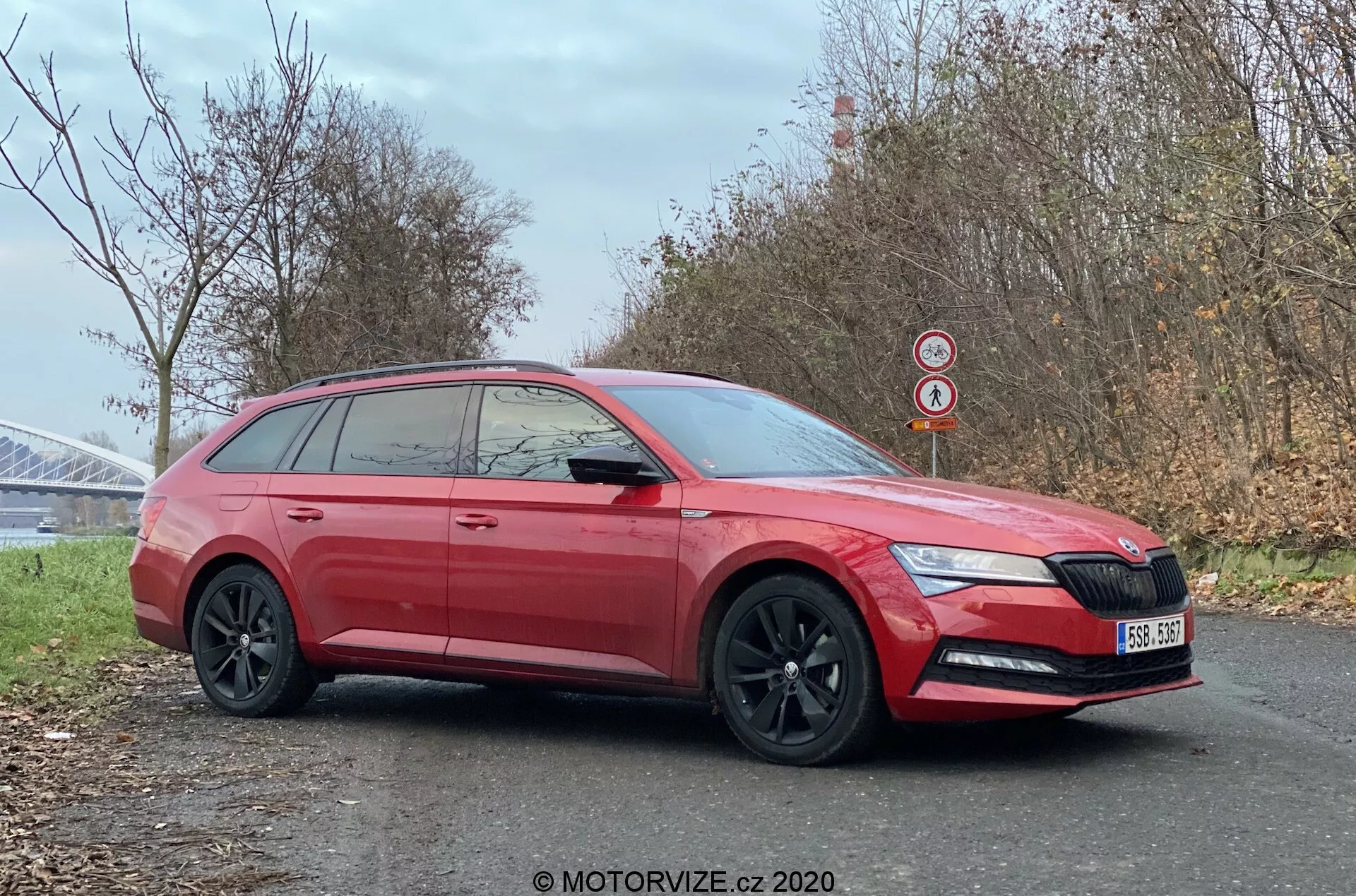 Profilo laterale di una Skoda Superb station wagon rossa del 2019 parcheggiata sul ciglio della strada, che mette in risalto il suo design moderno con fari anteriori affilati che includono luci diurne a LED, distintiva griglia frontale con badge logo, cerchi in lega neri, vetri oscurati per la privacy e una linea del tetto allungata tipica di una station wagon. La foto, incorniciata per enfatizzare sia il lato che la parte anteriore leggermente accorciata, cattura dettagli come indicatori di direzione integrati negli specchietti laterali, maniglie delle portiere in tinta carrozzeria, protezioni inferiori e uno spoiler sul tetto posteriore. L'immagine chiara scattata durante una stagione fredda vicino a una possibile struttura di ponte consente la visibilità delle caratteristiche dell'auto e dell'ambiente circostante.