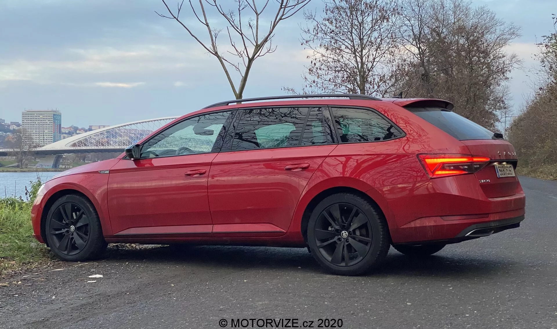 L'immagine presenta una moderna Skoda Superb 2019 di colore rosso vibrante, vista da un angolo posteriore di tre quarti. Parcheggiata su un'area pavimentata con un fiume e un ponte sullo sfondo, l'auto mostra linee eleganti, un tetto posteriore inclinato, finestrini leggermente oscurati con rifiniture cromate e uno spoiler posteriore integrato. Il design sportivo è evidenziato da grandi ruote in lega nere con pinze dei freni visibili, specchietti laterali neri a contrasto, maniglie delle porte a filo, luci posteriori a LED e sensori di parcheggio sul paraurti posteriore, indicando un alto livello di allestimento con caratteristiche di prestazioni e assistenza alla guida avanzate.
