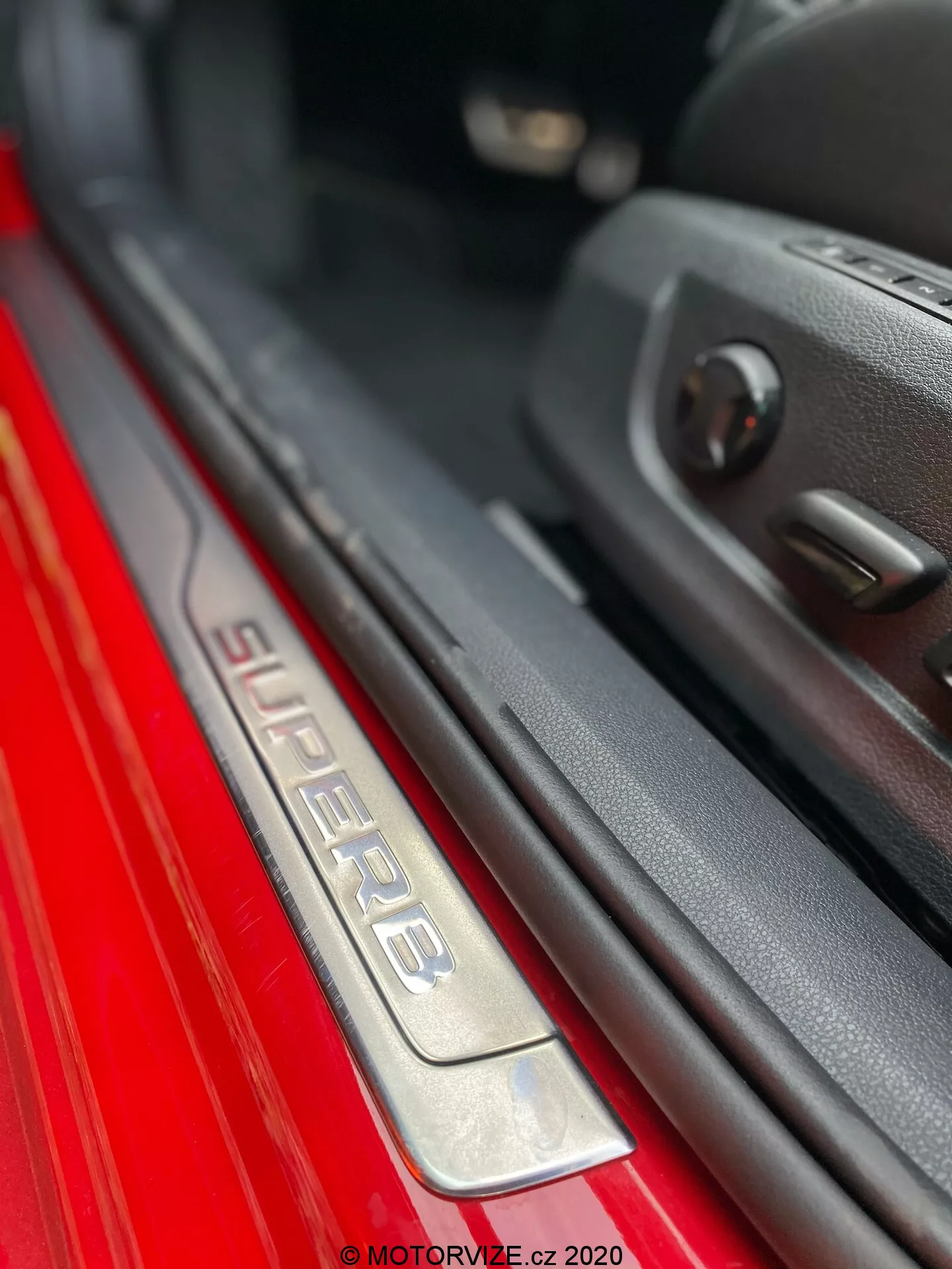 Vista ravvicinata della soglia della portiera lato passeggero della Skoda Superb 2019. L'immagine presenta un'angolazione inclinata che mostra l'iscrizione 'SUPERB' sulla soglia in acciaio inossidabile spazzolato, illuminata per un miglior effetto estetico. La vernice esterna rossa contrasta nettamente con l'interno scuro. Il bordo della porta con pannello in plastica e i comandi dei finestrini elettrici sono visibili, evidenziando caratteristiche avanzate. La ridotta profondità di campo mette in risalto la soglia, creando un effetto bokeh sullo sfondo.