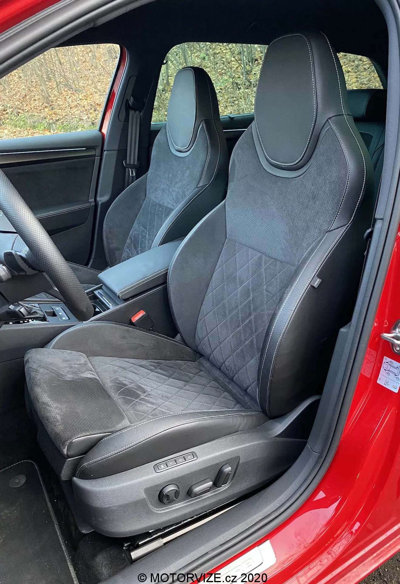 Interni della Škoda Superb 2019 focalizzati sul sedile del conducente. Il sedile sportivo a secchiello con rivestimento in pelle e materiale simile a camoscio, cuciture trapuntate, poggiatesta integrato, contorni pronunciati delle spalle e comandi di regolazione elettronici indicano un design orientato alle prestazioni, lusso e funzionalità avanzate di comfort. La cornice della porta visibile rossa suggerisce che l'esterno potrebbe essere rosso.