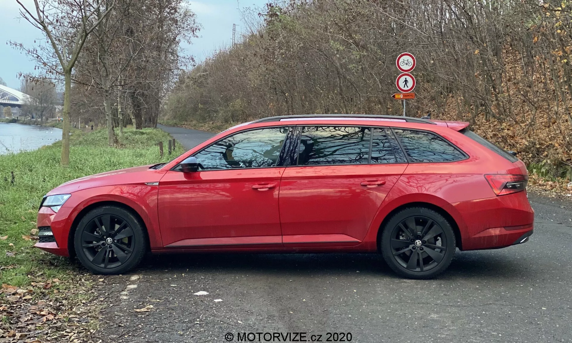 Immagine di profilo laterale di una moderna station wagon Skoda Superb 2019 (Superb-Limousine) di colore rosso, che mostra la linea del tetto allungata, le ruote in lega nera e gli specchietti laterali in tinta con gli indicatori di direzione integrati. L'auto è parcheggiata su un sentiero asfaltato accanto a una zona erbosa, con alberi e un corpo d'acqua sullo sfondo, alla luce naturale del giorno.