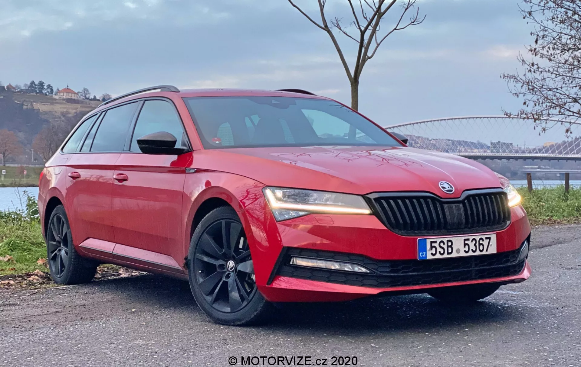 Vista del quarto frontale a bassa angolazione di una Škoda Superb rossa del 2019 parcheggiata all'aperto, che mette in risalto le linee di design, la griglia, le ruote, il profilo laterale e le leghe scure sportive della wagon sullo sfondo di un paesaggio pittoresco con acqua e alberi.