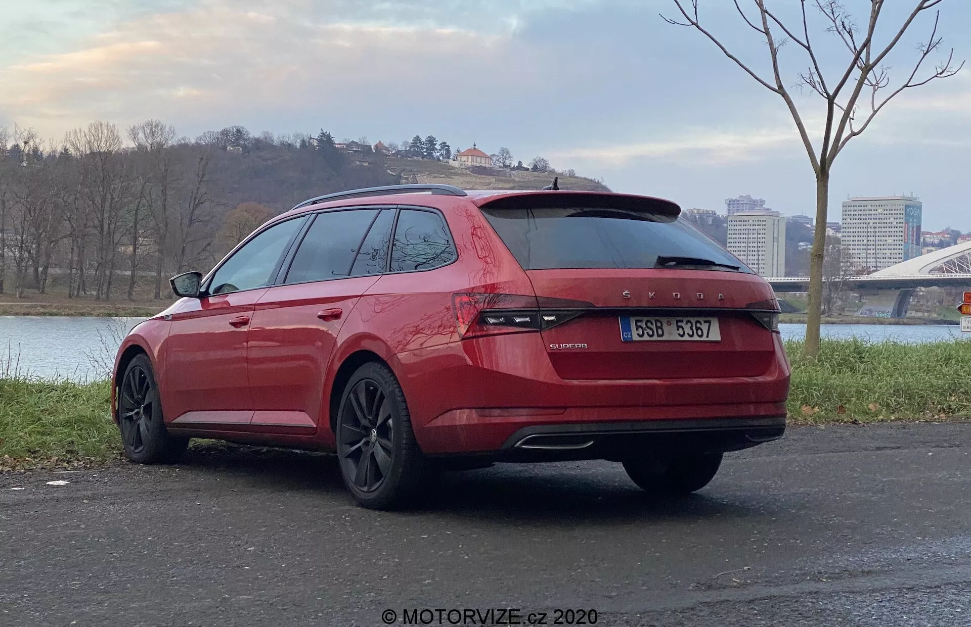 Vista posteriore angolata di una station wagon Skoda Superb 2019 rossa parcheggiata all'aperto, focalizzandosi sul retro e sul lato passeggero del veicolo. Con luci posteriori continue lungo il portellone, doppi tubi di scarico, vetri oscurati, cerchi in lega dal design scuro e situata su una superficie pavimentata con uno specchio d'acqua e un ponte sullo sfondo.