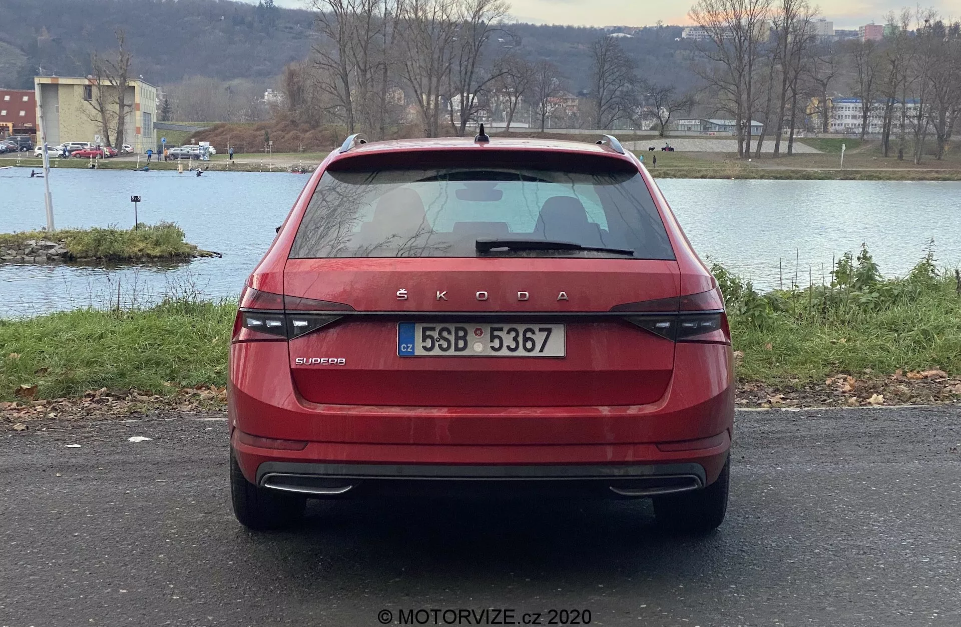 Vista posteriore di una Škoda Superb 2019 limousine rossa, con il distintivo marchio Škoda e l'iscrizione del modello OCTAVIA sul cofano. L'immagine mette in risalto le moderne luci posteriori orizzontali, la targa centrale, il tergicristallo posteriore, il lunotto posteriore oscurato con linee disappannanti, la luce di stop montata in alto, i sensori di parcheggio nel paraurti e i riflettori. L'auto è parcheggiata all'aperto con un corpo d'acqua, alberi e un orizzonte sullo sfondo.