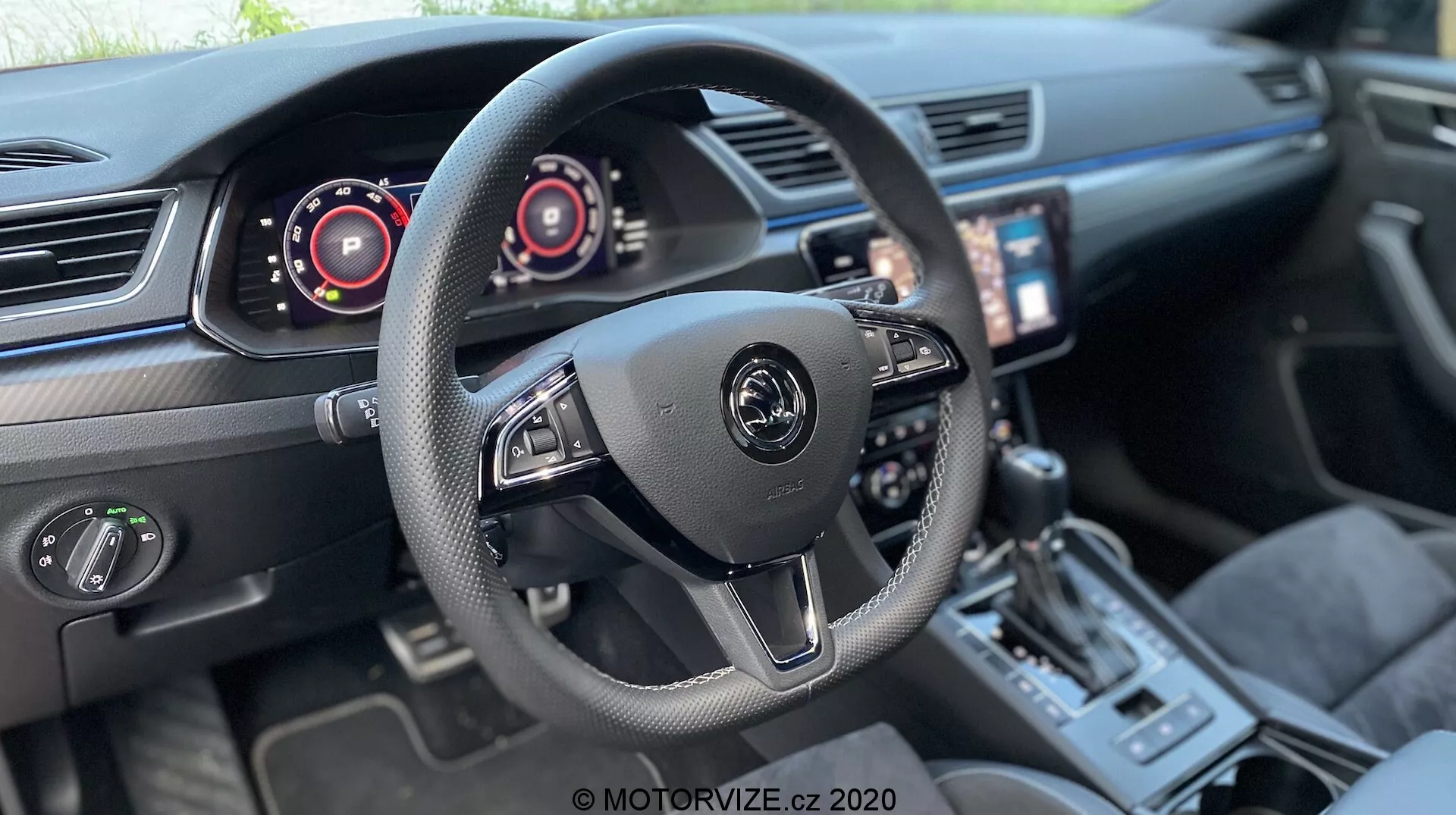 Vista dell'interno di una Škoda Superb Limousine 2019, focalizzata sull'area del posto di guida. Volante centrale a tre razze con logo del produttore e comandi per audio e sistemi di bordo, inclusi paddles dietro al volante. Quadri strumenti digitali alle spalle, indicanti tachimetro e contagiri. Schermo touchscreen per l'infotainment orientato verso il conducente, climatizzatore bizona, leva del cambio sportiva, selettore delle modalità di guida e sedili in pelle di qualità con cuciture a contrasto, suggeriscono un veicolo moderno e di alta gamma con tecnologia avanzata.