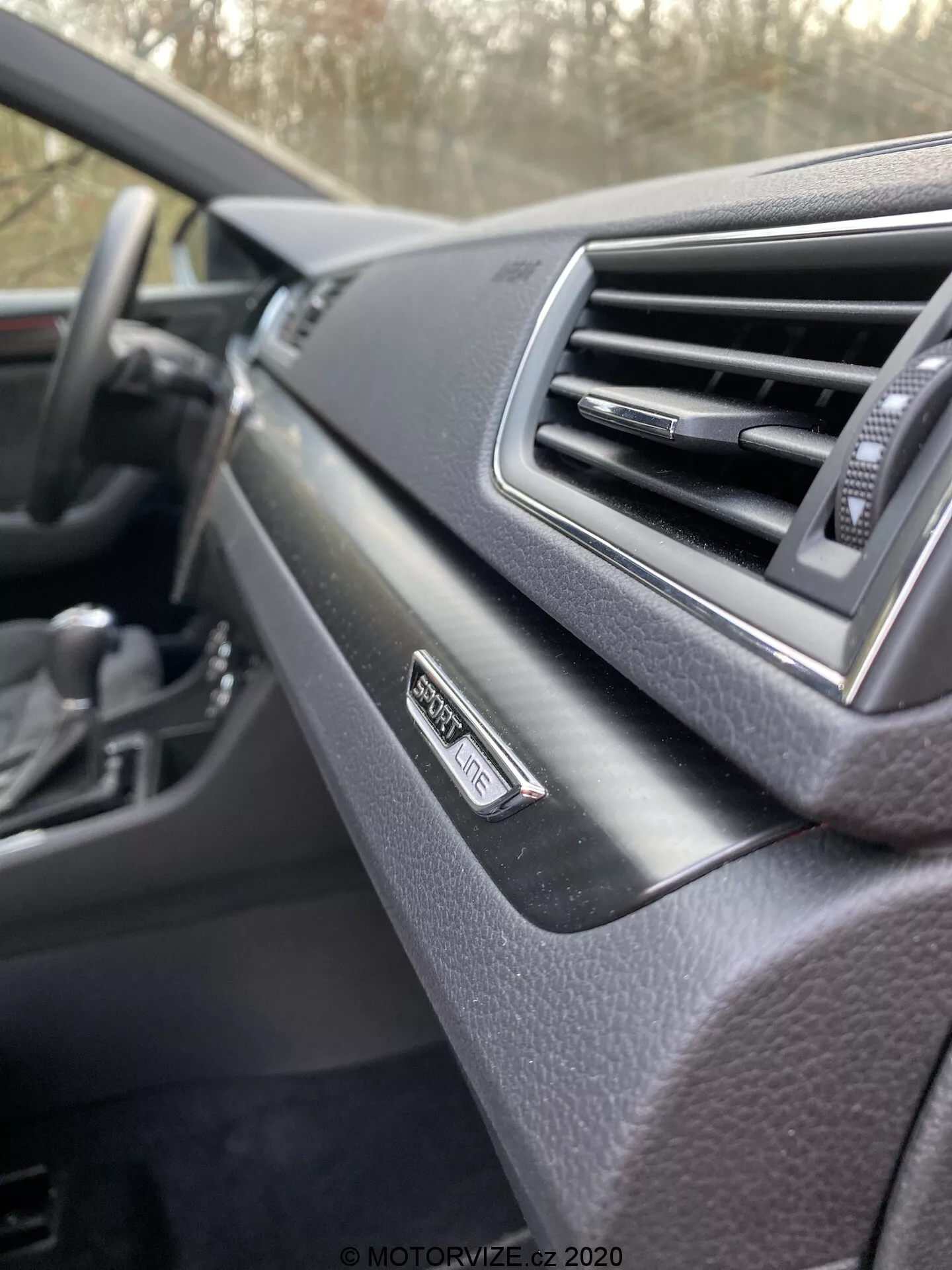 Primo piano degli interni della Škoda Superb 2019, cruscotto dalla prospettiva del passeggero. Messa a fuoco sul badge 'Superb' vicino alla bocchetta d'aria, design moderno con finitura morbida al tatto. Volante sfocato sullo sfondo, schema di colore scuro.