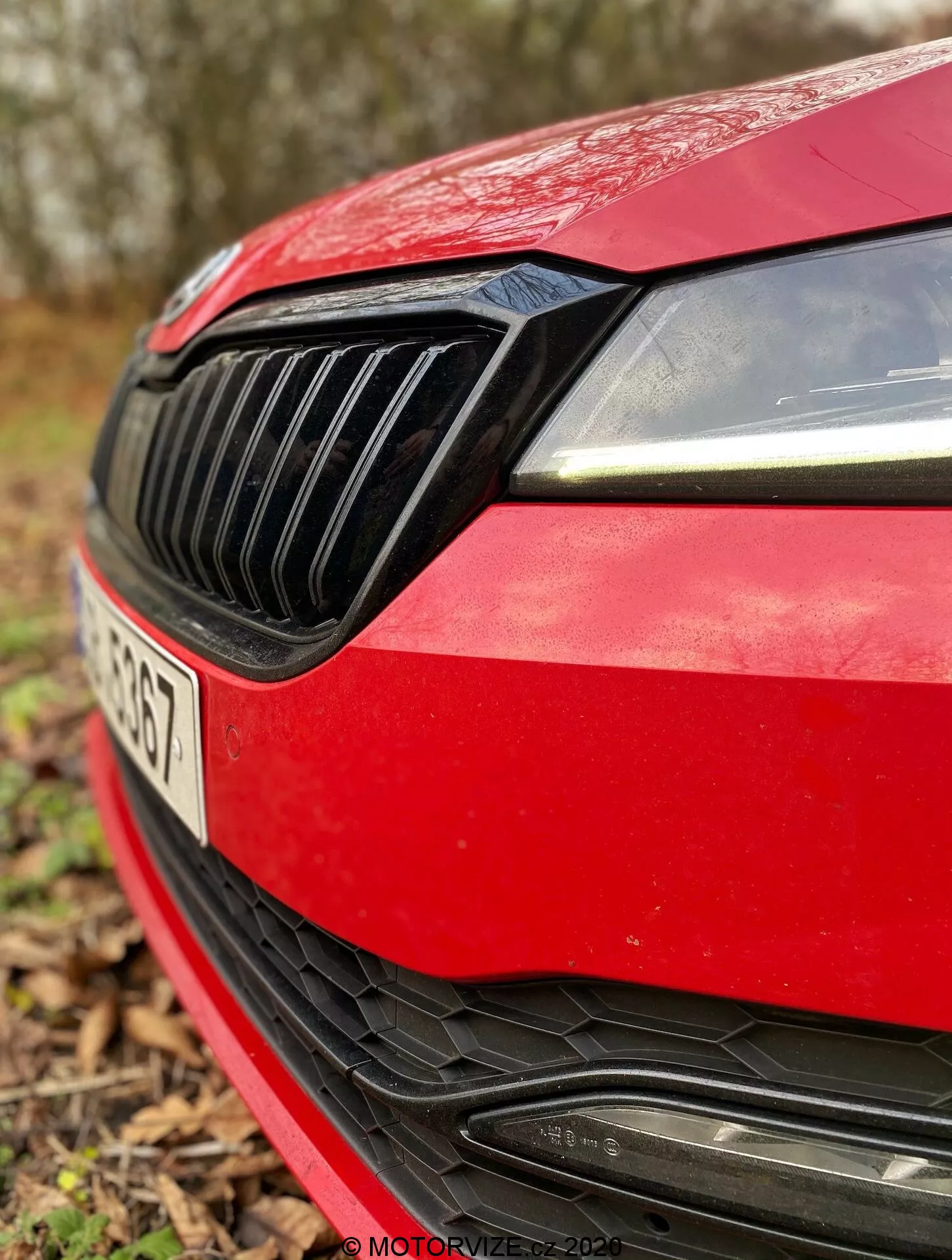 Primo piano del quarto anteriore sinistro di una Skoda Superb Limousine 2019 rossa. L'immagine cattura il design moderno del faro destro con luci diurne a LED, la griglia nera con listelli verticali, il bordo aerodinamico del cofano rosso, il paraurti anteriore sportivo con una grande presa d'aria con listelli orizzontali e lo spazio per il distintivo del produttore, parzialmente nascosto. La targa è leggermente visibile ma fuori fuoco a causa della poca profondità di campo, con uno sfondo naturale sfocato.