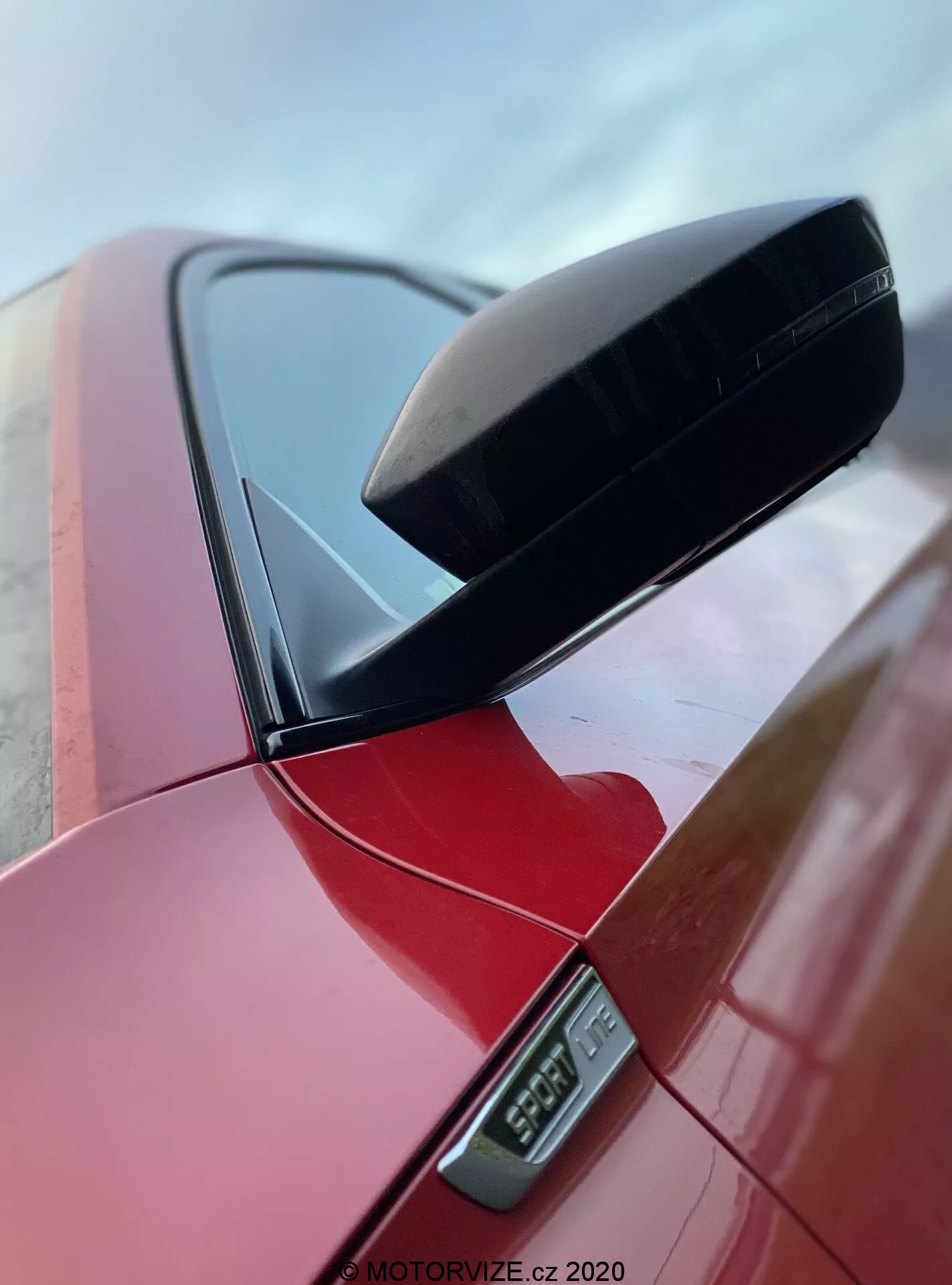 Vista ravvicinata dello specchietto retrovisore laterale su una Skoda Superb 2019 berlina di color rosso vibrante, evidenziando la calotta aerodinamica nera dello specchio e la lampada laterale a LED sottostante, con una parte del pilastro dell'auto visibile, ripresa da un'angolazione bassa che enfatizza il design pulito e la finitura lucida, contrassegnata da una filigrana MOTORVIZE.cz 2020.