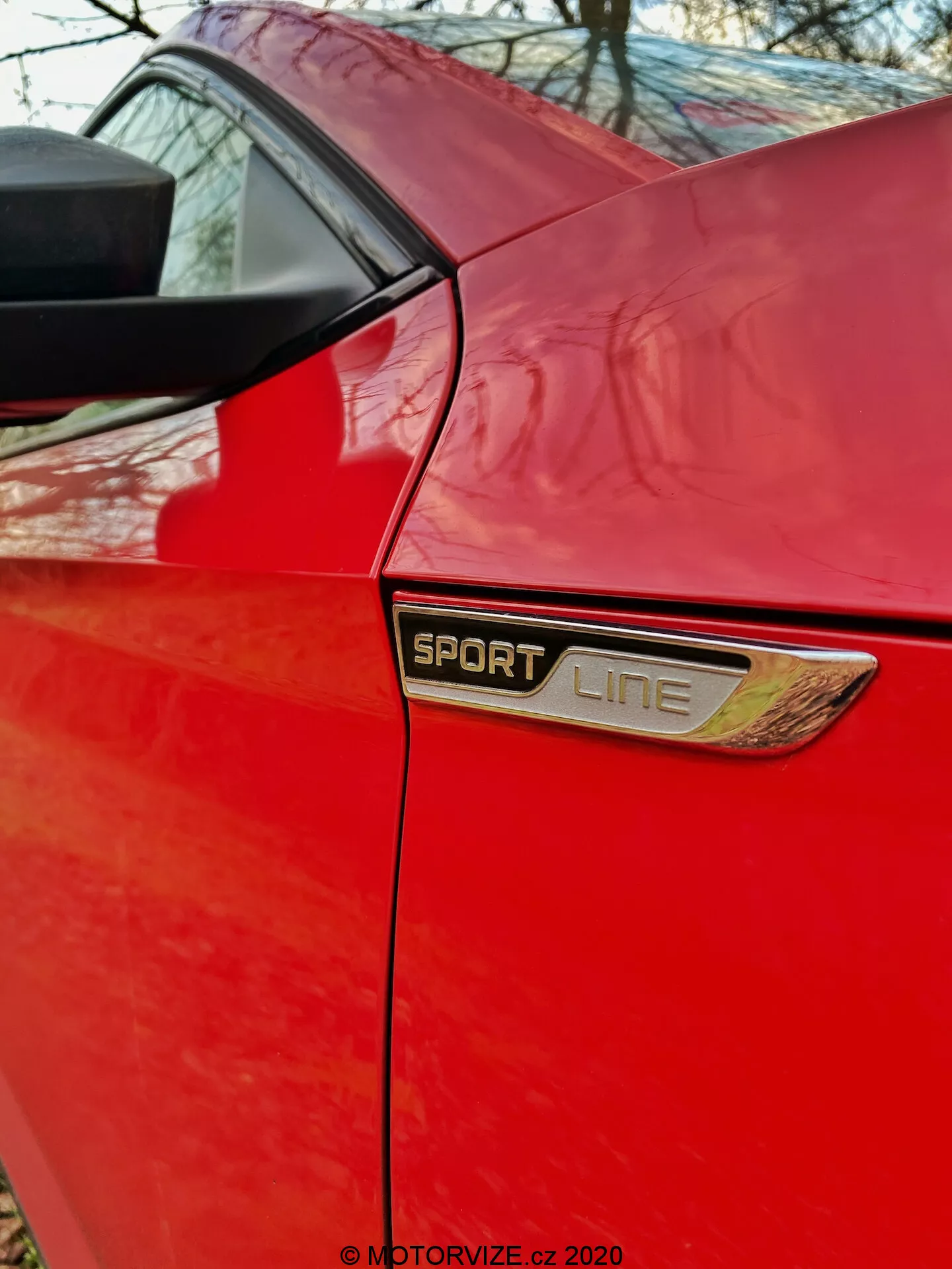 Vista ravvicinata e angolata del pannello posteriore di una Škoda Superb 2019 limousine rossa. L'immagine si concentra sull'area dietro la porta laterale, mettendo in evidenza il distintivo 'SPORT LINE' centrato nel fotogramma con una cornice cromata o metallica e una decorazione ventilata aggiuntiva sotto. Sopra il distintivo, è visibile il bordo inferiore della finestra laterale posteriore e il montante posteriore che sostiene la struttura del tetto. La foto mette in risalto i livelli di allestimento distintivi, le caratteristiche estetiche e la qualità della verniciatura della versione sportiva