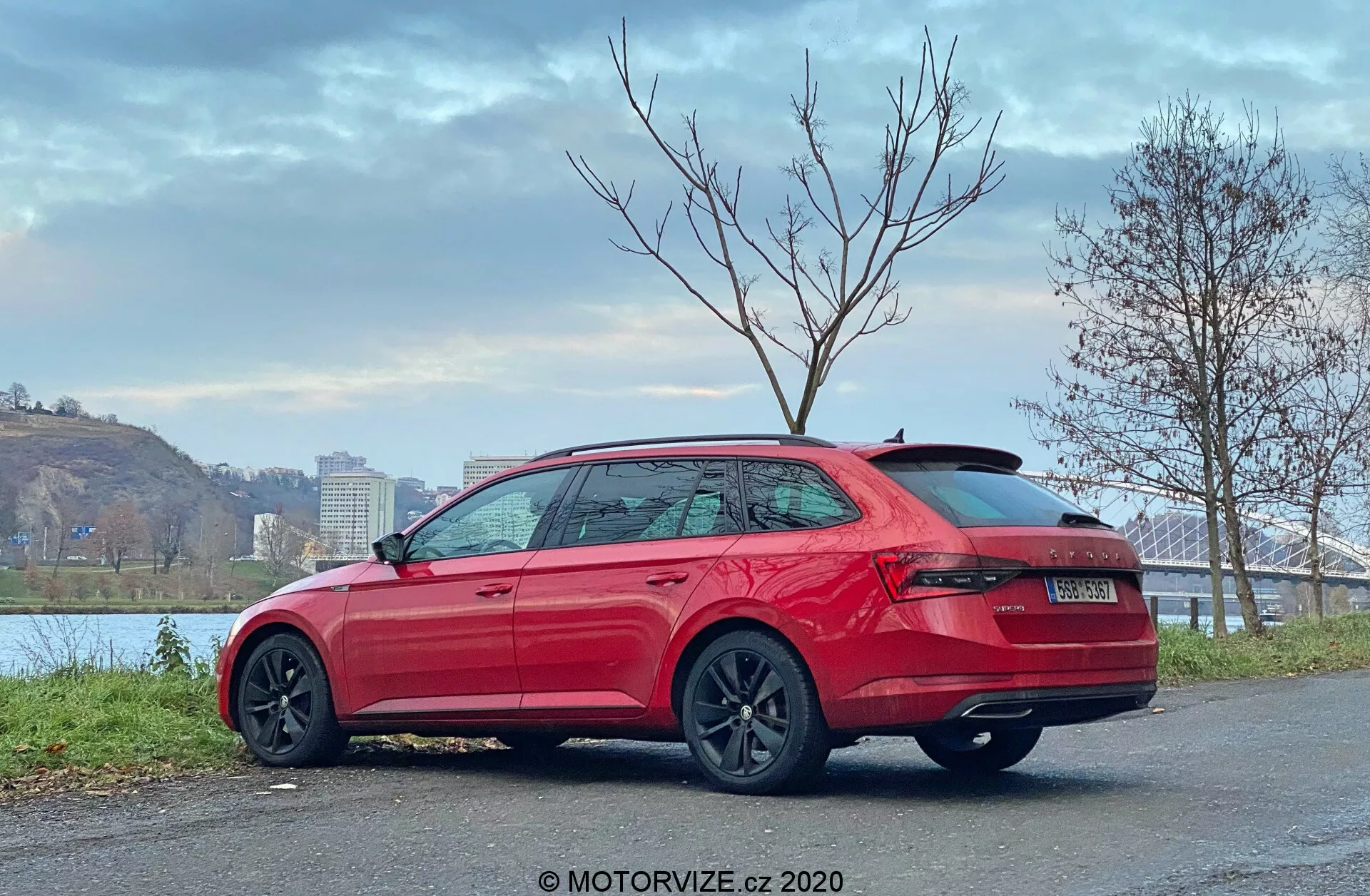 Immagine di una Skoda Superb 2019, station wagon, vista posteriore a tre quarti, che mostra fanali posteriori avvolgenti montati in alto, paraurti robusto, nessun tubo di scarico visibile, spoiler sul tetto, linee eleganti del contorno laterale, maniglie delle porte in tinta con la carrozzeria, protezioni sulle minigonne laterali, rifiniture dei finestrini cromate, ruote multi-razze scure, parcheggiata su una superficie pavimentata vicino alla riva del fiume con ponte e alberi, cielo nuvoloso, filigrana MOTORVIZE.cz 2020.