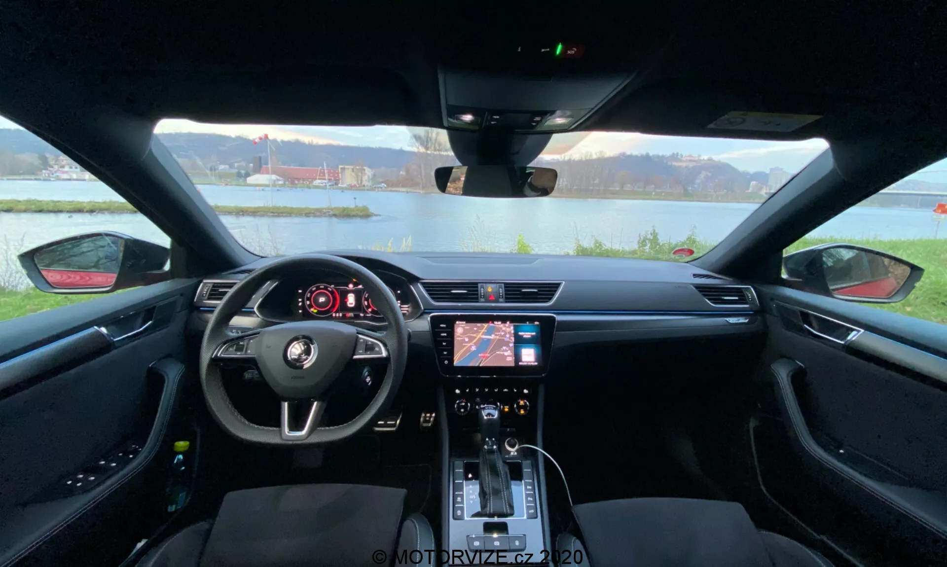 Vista interna di una Skoda Superb 2019 dal sedile posteriore in direzione della plancia, che cattura l'intera larghezza dell’interno dell’auto. Il volante centrale con comandi integrati e il logo Skoda è visibile in primo piano. Dietro al volante è presente un display digitale che mostra informazioni essenziali del veicolo. La console centrale dispone di un sistema di infotainment con navigazione e impostazioni multimediali, insieme ai controlli del clima e ai pulsanti delle funzioni sottostanti. La leva del cambio, i portabicchieri e i sedili dettagliati con cuciture a contrasto enfatizzano l'estetica moderna e il focus sul comfort e la convenienza del conducente.