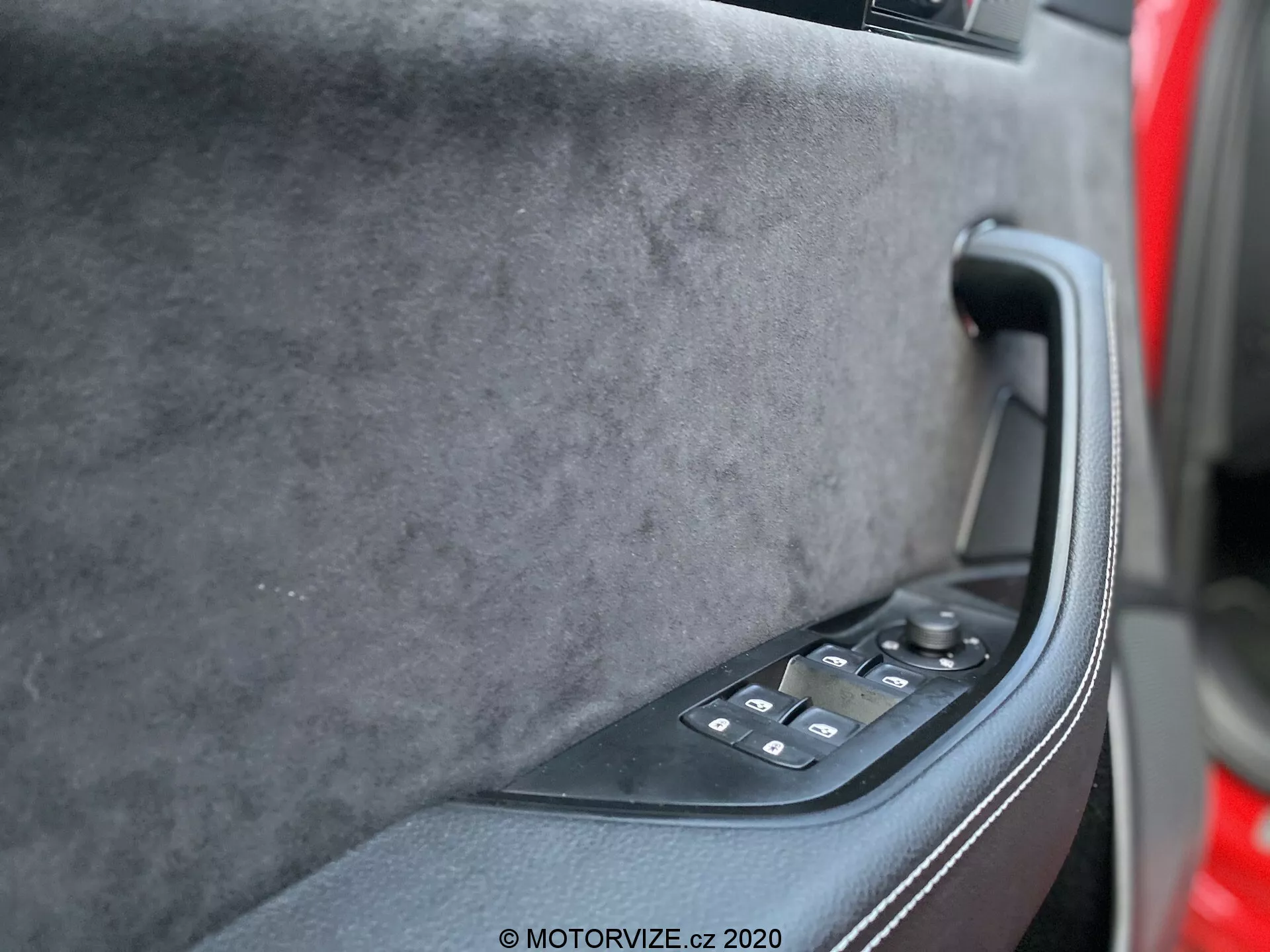 Il pannello interno della porta della Skoda Superb 2019 dalla prospettiva del sedile del conducente, evidenziando la sezione superiore soft-touch in tessuto premium, una maniglia ergonomica nera solida e un pannello di controllo nero lucido con regolazioni dei finestrini elettrici e possibilmente controlli degli specchietti elettrici. Toni scuri e smorzati, tecnologia moderna e un design lussuoso sono evidenti.