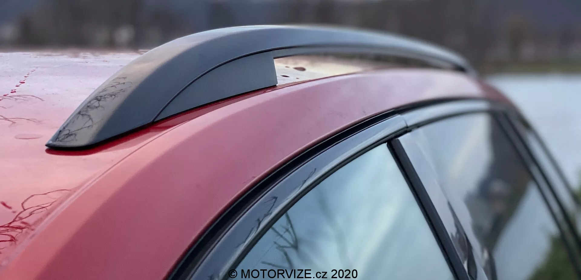 Vista laterale ravvicinata di una sezione del veicolo Škoda Superb 2019, concentrandosi sulla linea del tetto rosso scuro, il montante laterale e l'accessorio dell'antenna a pinna di squalo, con gocce d'acqua visibili che indicano una superficie cerata o idrofoba; il design aerodinamico dell'auto è evidente attraverso la curvatura e l'inclinazione del tetto