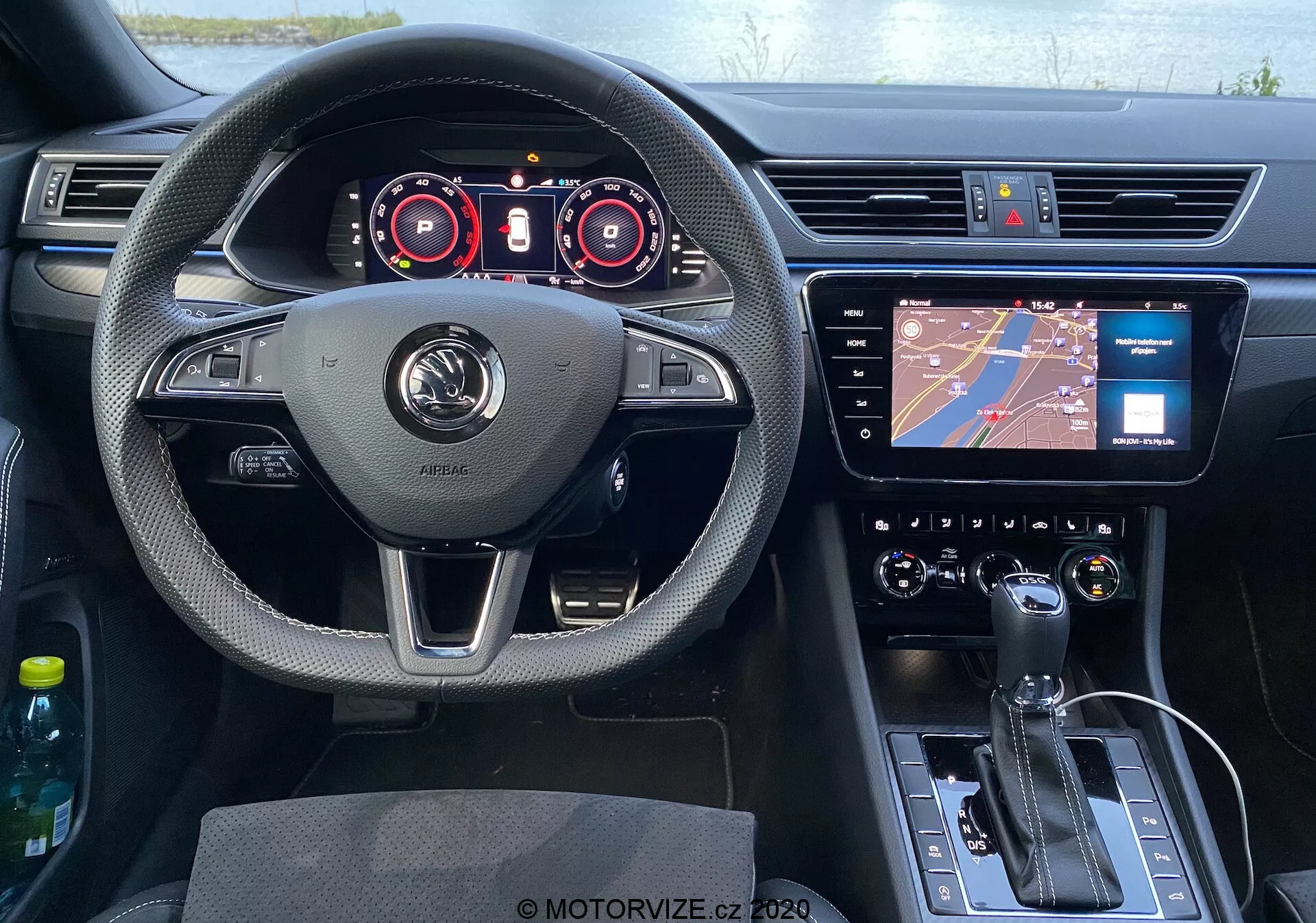 Vista dal sedile del conducente dell'abitacolo della Skoda Superb 2019 con un volante centrato dotato di poggia pollici, pulsanti multifunzione e logo del produttore ben visibile. Dietro il volante è presente un quadro strumenti digitale con indicatori per tachimetro e contagiri, insieme a display aggiuntivi per dettagli operativi. Al centro del cruscotto si trova un touchscreen di infotainment con navigazione, e sotto di esso un sistema di controllo del clima con pulsanti e manopole. La console centrale inferiore include la leva del cambio per la trasmissione automatica, pulsante del freno di stazionamento elettronico e una manopola di controllo. Sono visibili la portiera del conducente e lo specchietto retrovisore, con i comandi della porta evidenti. L'abitacolo sfoggia una combinazione di materiali testurizzati, accenti cromati, volante rivestito in pelle e plastica nera lucida sulla console, conferendo un'estetica moderna.