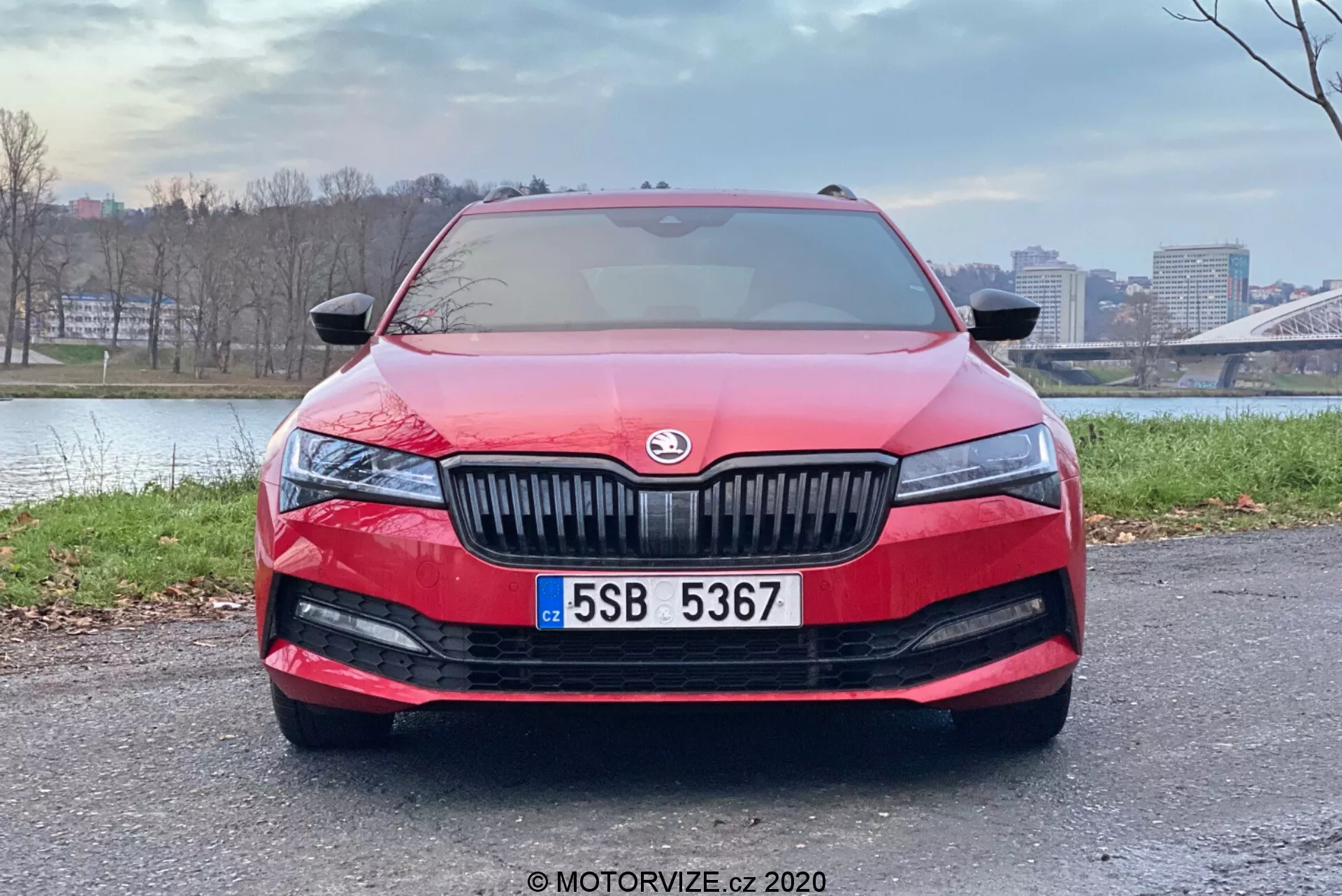 Vista frontale di una moderna Skoda Superb Limousine 2019 rossa, che mostra i suoi lisci contorni aerodinamici, la griglia prominente con alette verticali con il logo del produttore al centro, i gruppi ottici a LED slanciati ed allungati, l'ampia griglia di aspirazione dell'aria sul paraurti inferiore con fari fendinebbia, la targa centrale e possibilmente un sensore o una telecamera dei sistemi di assistenza avanzata alla guida. Il parabrezza riflette il cielo, suggerendo sensori pioggia per i tergicristalli o un sensore di luce per l'attivazione automatica dei fari. L'auto è parcheggiata in un'area pavimentata con uno sfondo urbano, mettendo in risalto il suo design e le caratteristiche.