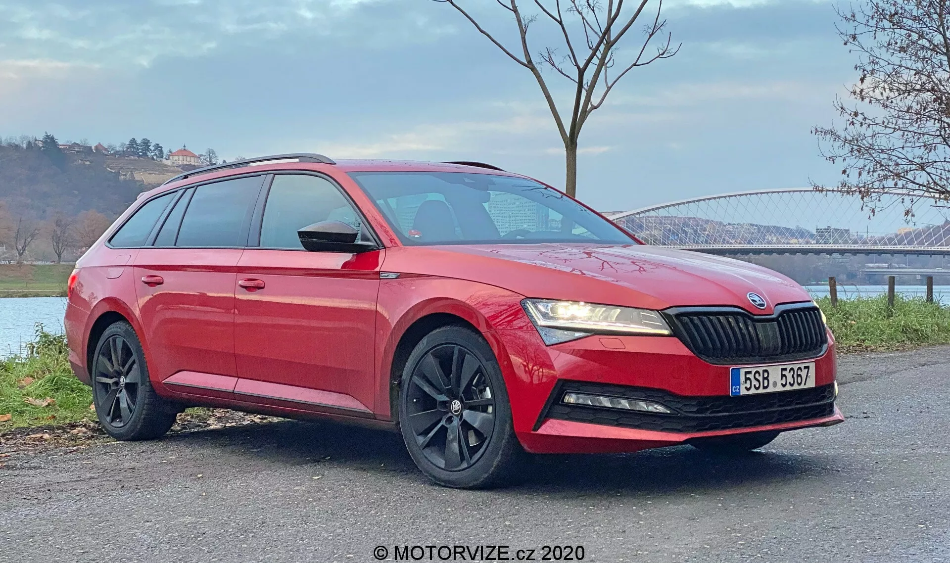 Immagine di una station wagon Skoda Superb 2019 rossa catturata da una vista frontale a tre quarti. La foto mette in evidenza la griglia frontale con lamelle verticali, fari angolari eleganti, fendinebbia nell'area del paraurti inferiore, cerchi in lega neri a più razze e carrozzeria lucida con linee distintive e barre sul tetto. Sono visibili anche gli specchietti laterali in tinta con la carrozzeria con indicatori di direzione integrati e i vetri posteriori oscurati. Il veicolo, posizionato su una superficie piana all'aperto, è visto con uno sfondo naturale che presenta un ponte in lontananza e un corpo idrico. Le condizioni di luce diurna enfatizzano sia le sezioni inferiori che superiori dell'auto.