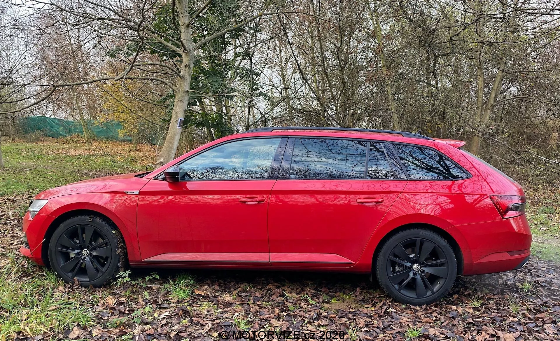 Skoda Superb station wagon del 2019 in rosso brillante, vista di profilo laterale. Caratteristiche di design moderno includono linee geometriche nitide, cerchi in lega neri a più razze, vetri oscurati con montanti B neri e una linea del tetto inclinata per un profilo aerodinamico elegante. L'auto è parcheggiata su un'area erbosa accanto a un sentiero, con la natura sullo sfondo, suggerendo un'adeguatezza per le uscite familiari o le avventure.
