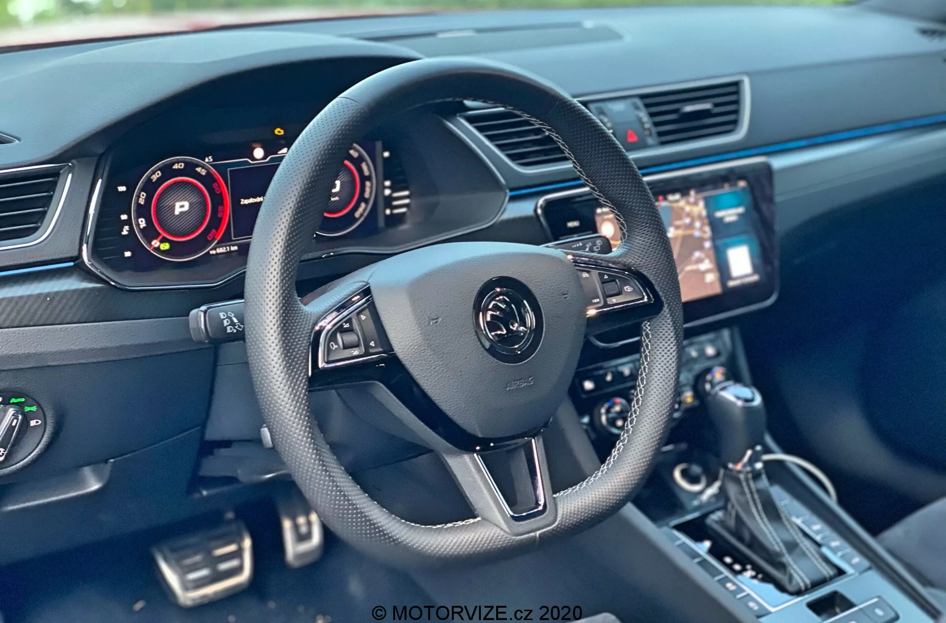 Interni della Skoda Superb 2019 dalla prospettiva del sedile del conducente, con un volante moderno a tre razze con comandi multifunzionali e un emblema centrale. Componenti visibili includono un cruscotto digitale, sistema di infotainment centrale con touch screen, console, leva del cambio, pulsanti e sistema di controllo del clima. La tecnologia degli interni dell'auto mostra elementi come cruscotti digitali e controlli del volante.