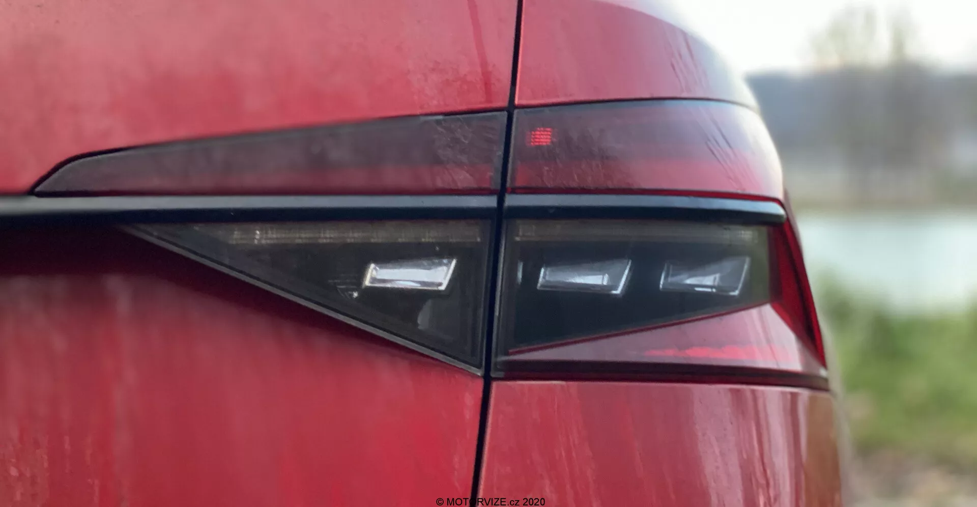 Dettaglio della parte posteriore di una Skoda Superb 2019, con particolare attenzione al gruppo ottico posteriore visto da un angolo laterale. Mostra un design moderno ed elegante con elementi luminosi a LED orizzontali racchiusi in una lente rosso scuro, riflettendo l'estetica contemporanea di veicoli sportivi o di lusso. La carrozzeria lucida e il listello orizzontale nero aumentano l'appeal visivo su uno sfondo sfocato.